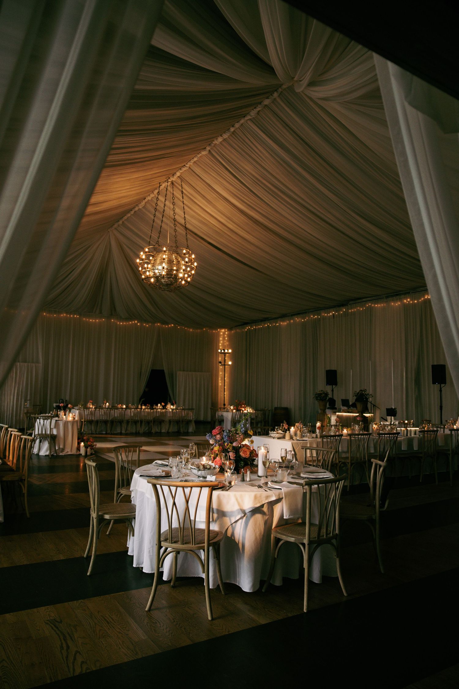Elegant wedding reception tent with draped white fabric ceiling, chandelier, round tables, and floral centerpieces in warm candlelight.
