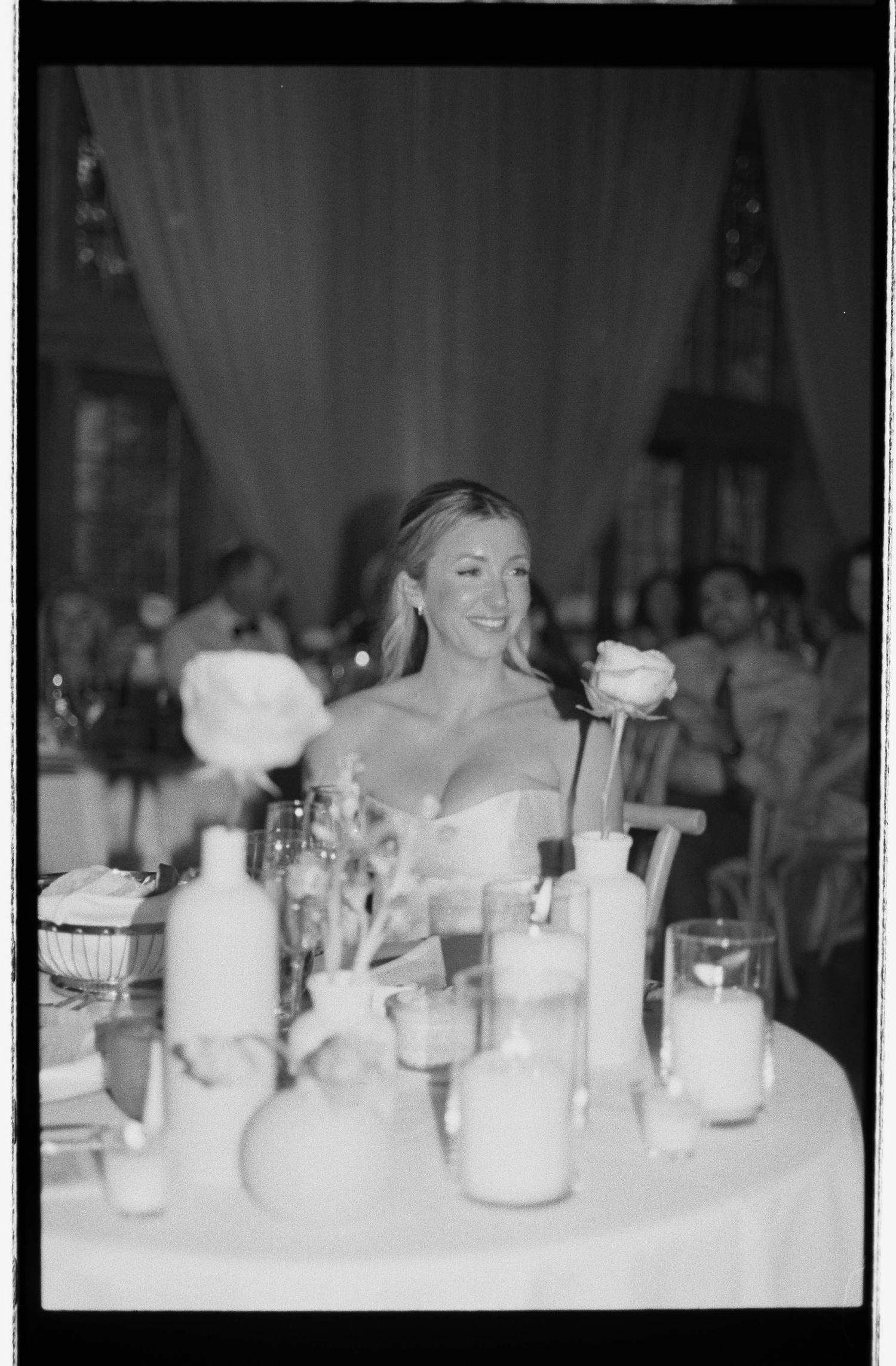 Black and white film photo of smiling bride at candlelit wedding reception table with flowers.