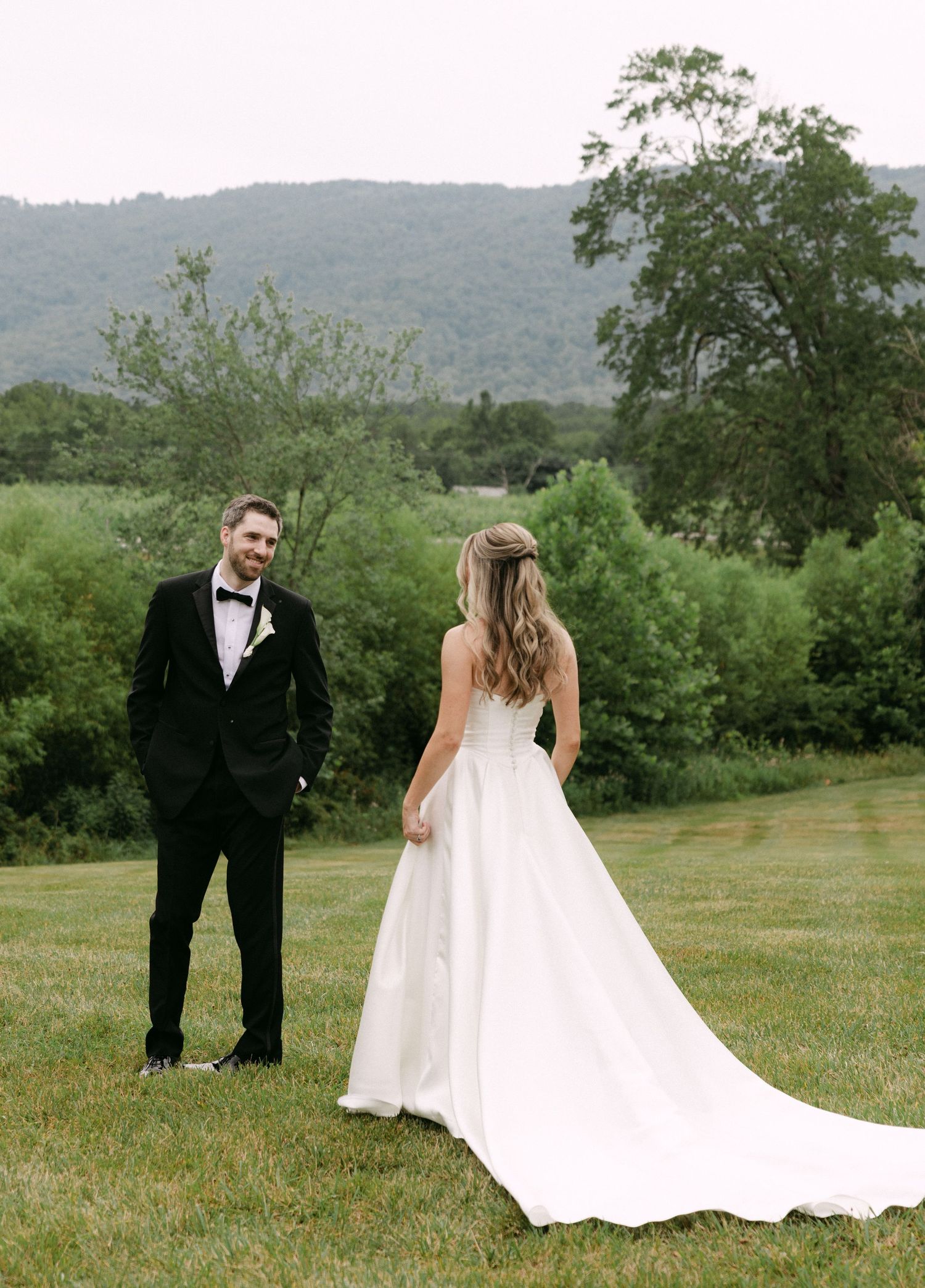 A bride in a white ball gown and groom in a black tuxedo share a first look on a lush green lawn with mountain views.