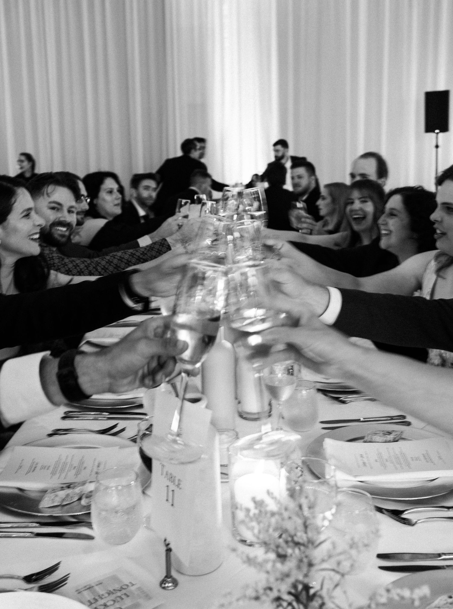 Black and white photo of wedding guests raising glasses in a celebratory toast at a reception dinner.