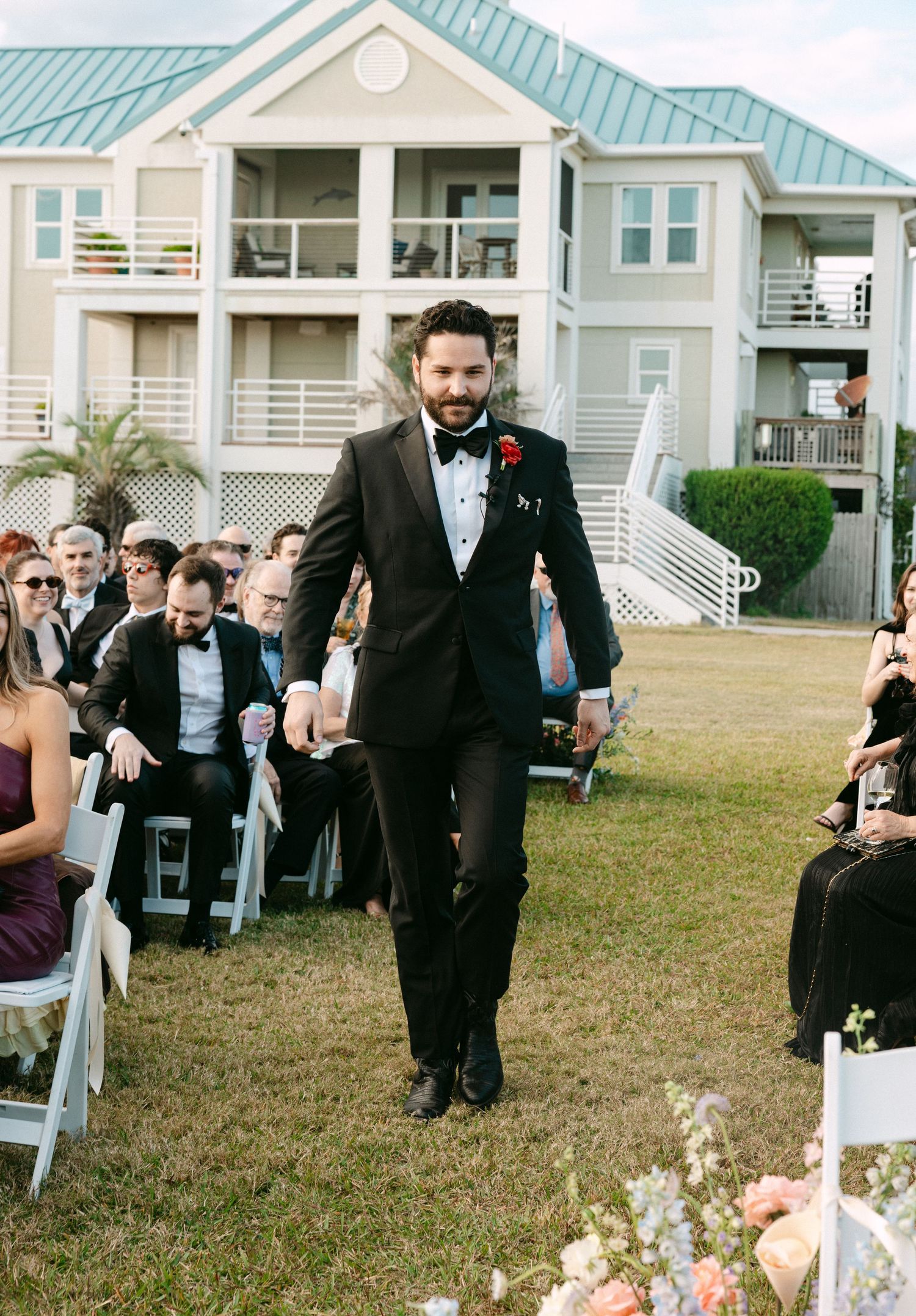 Groom in black tuxedo walks down outdoor wedding aisle with guests seated in white chairs.