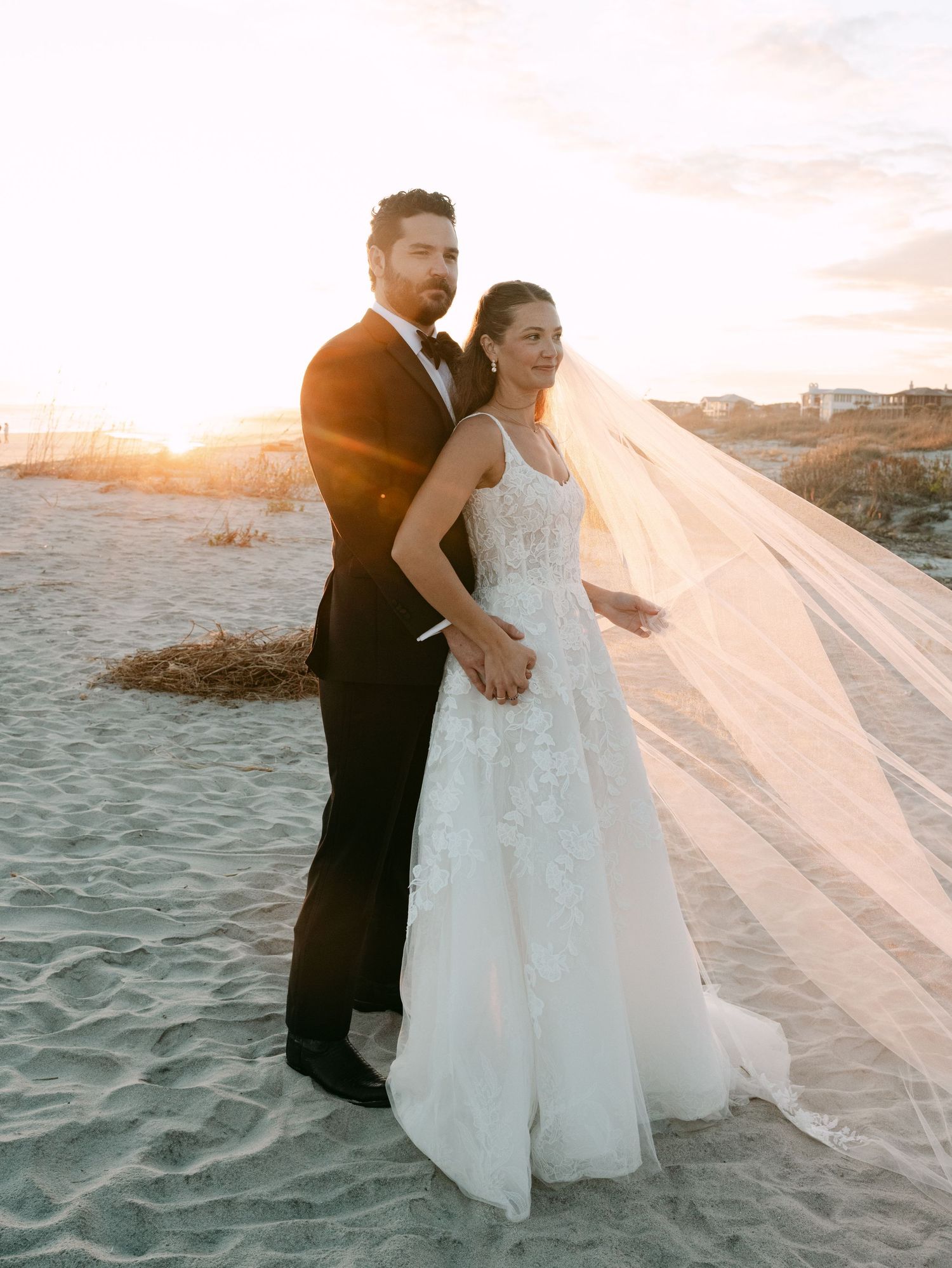 Wedding couple posing on beach at golden hour, bride wearing lace gown with flowing veil.