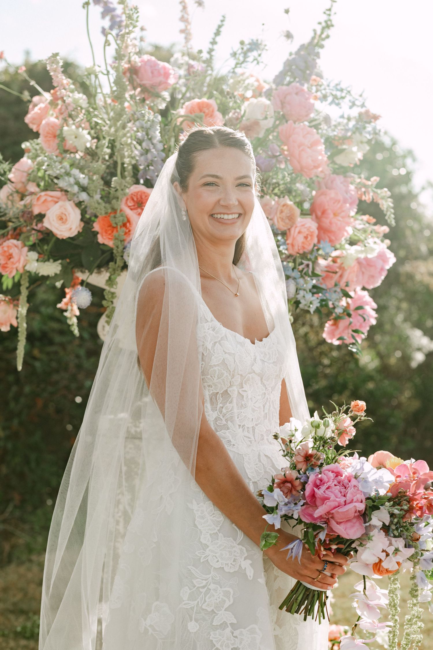 Smiling bride in lace wedding dress and veil holding pink bouquet in front of colorful floral arch.