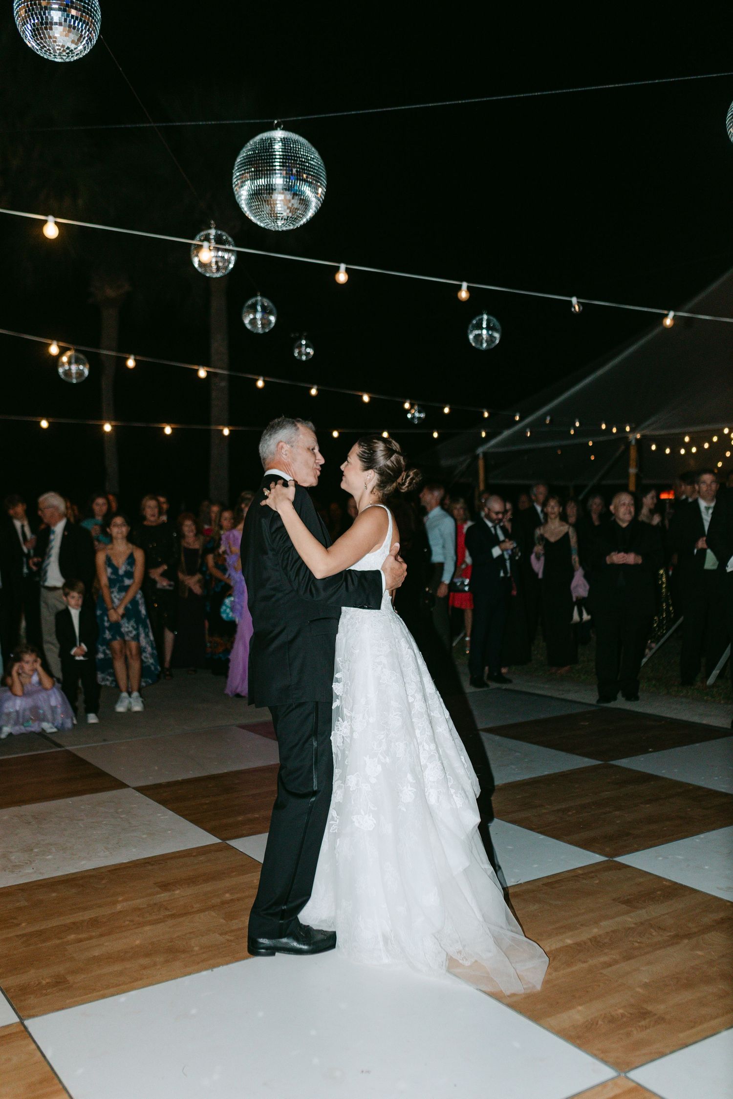 Bride and father share first dance under disco balls and string lights on checkered dance floor at night.