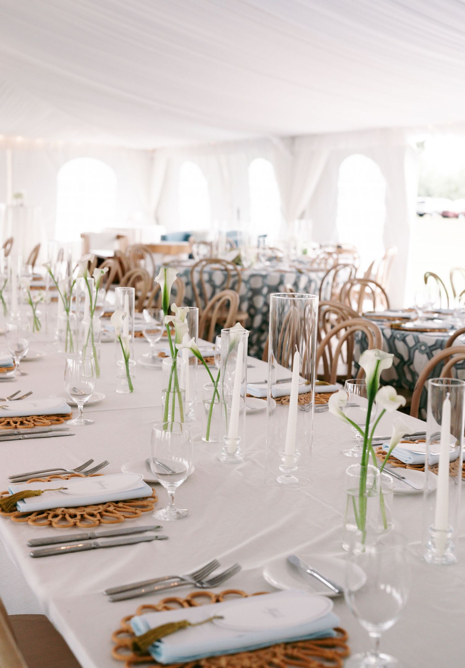 Elegant white wedding reception tables set with calla lilies in bud vases, taper candles, and wicker chargers.