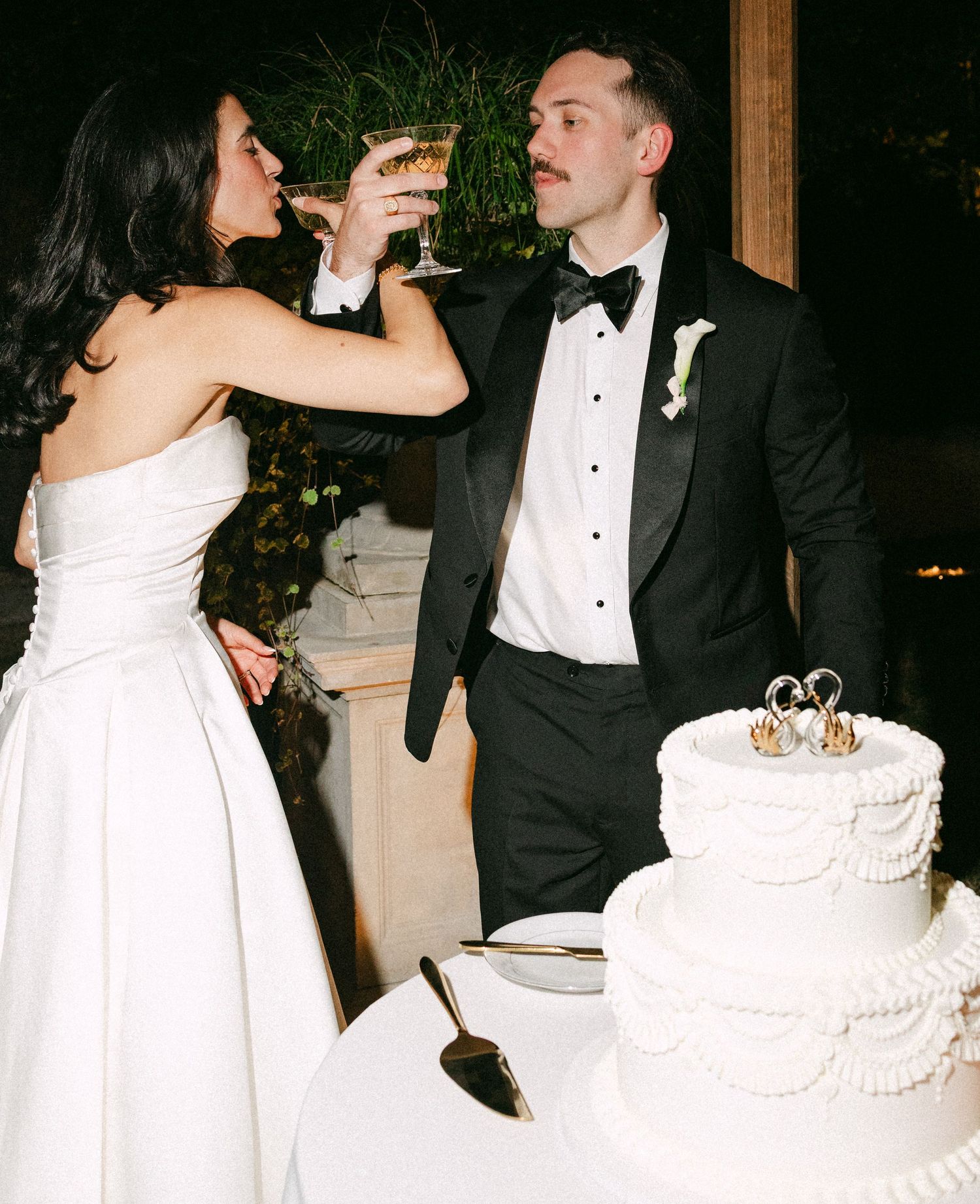 Bride and groom toast with champagne beside white tiered wedding cake at evening reception.