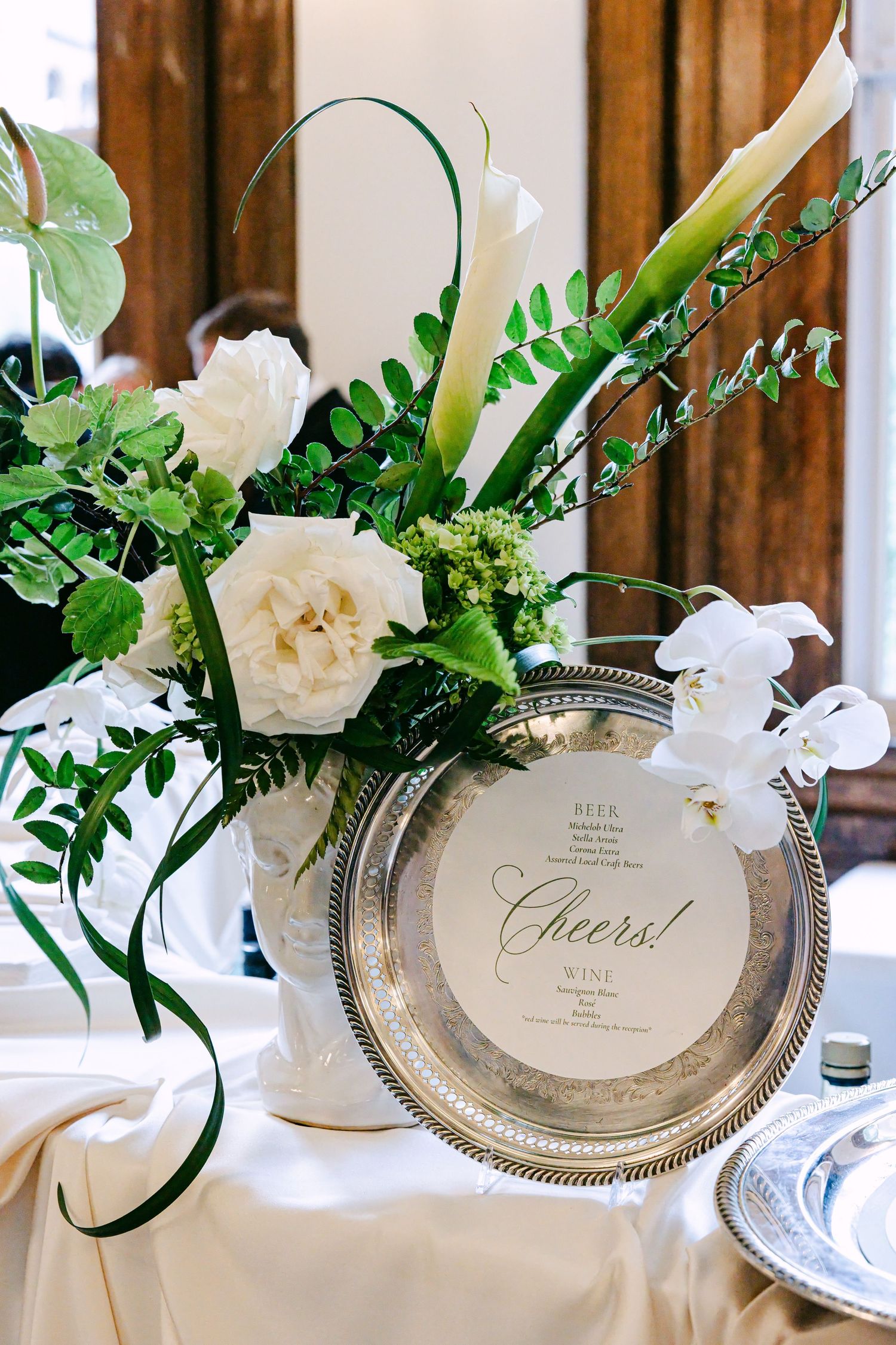 White wedding floral centerpiece with calla lilies beside a silver Cheers menu sign.