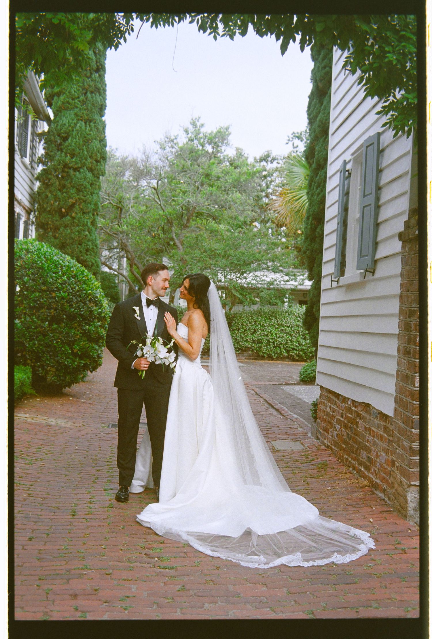 Bride and groom share a romantic moment on a brick pathway surrounded by lush greenery on their wedding day.