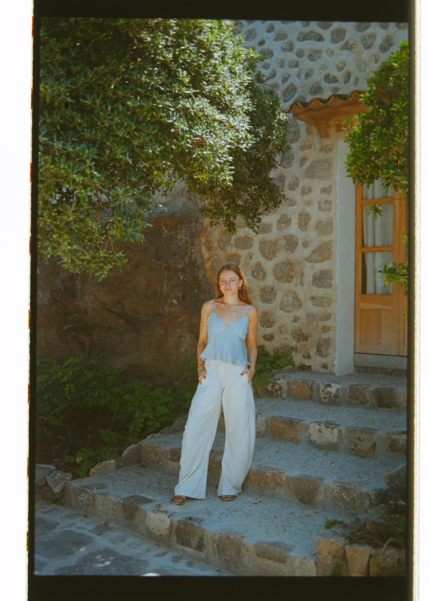 Woman in light blue top and white wide-leg pants standing on stone steps outside a rustic building with lush greenery.