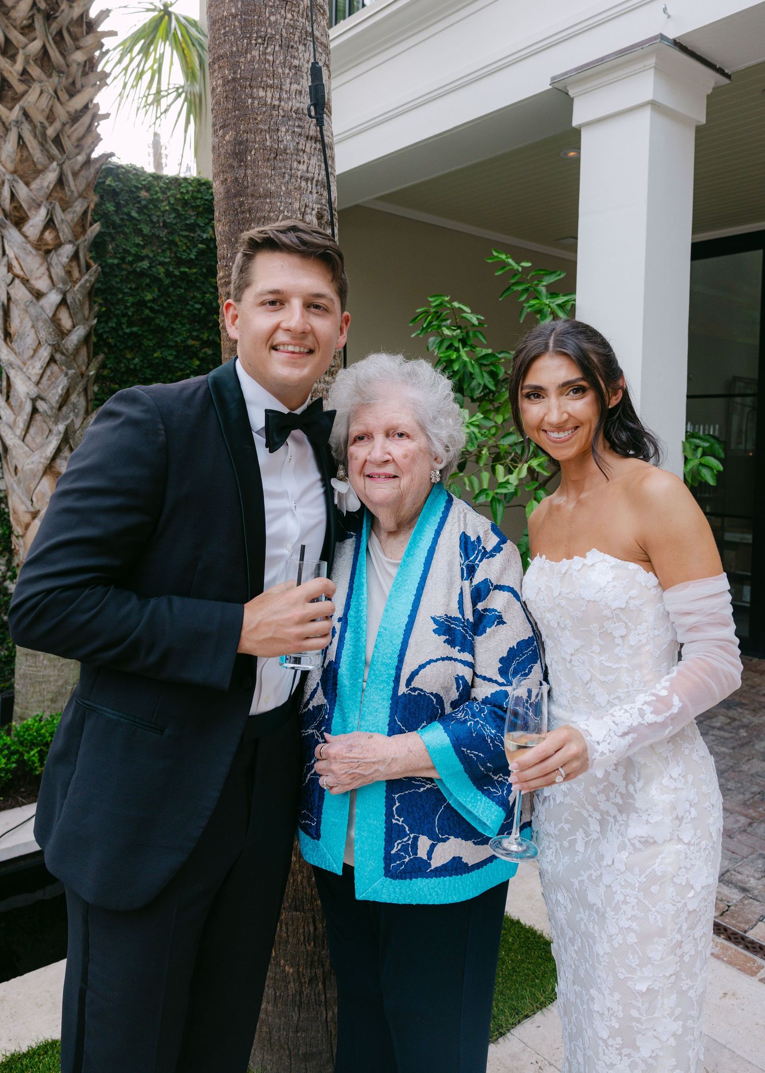 Formal wedding photo under palm trees with guests in evening attire.