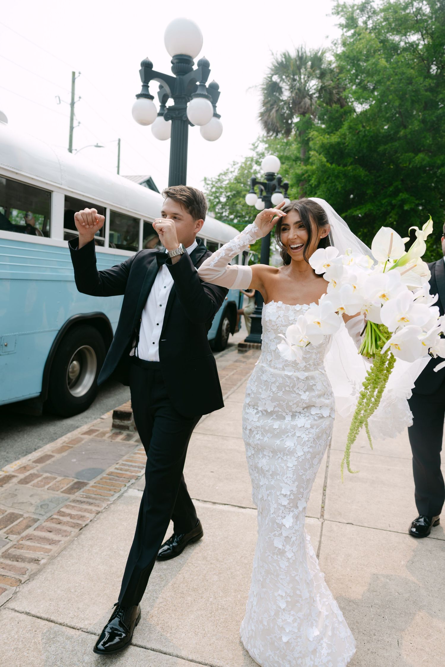 Bride in white dress and groom in tuxedo dance playfully on sidewalk outside venue.