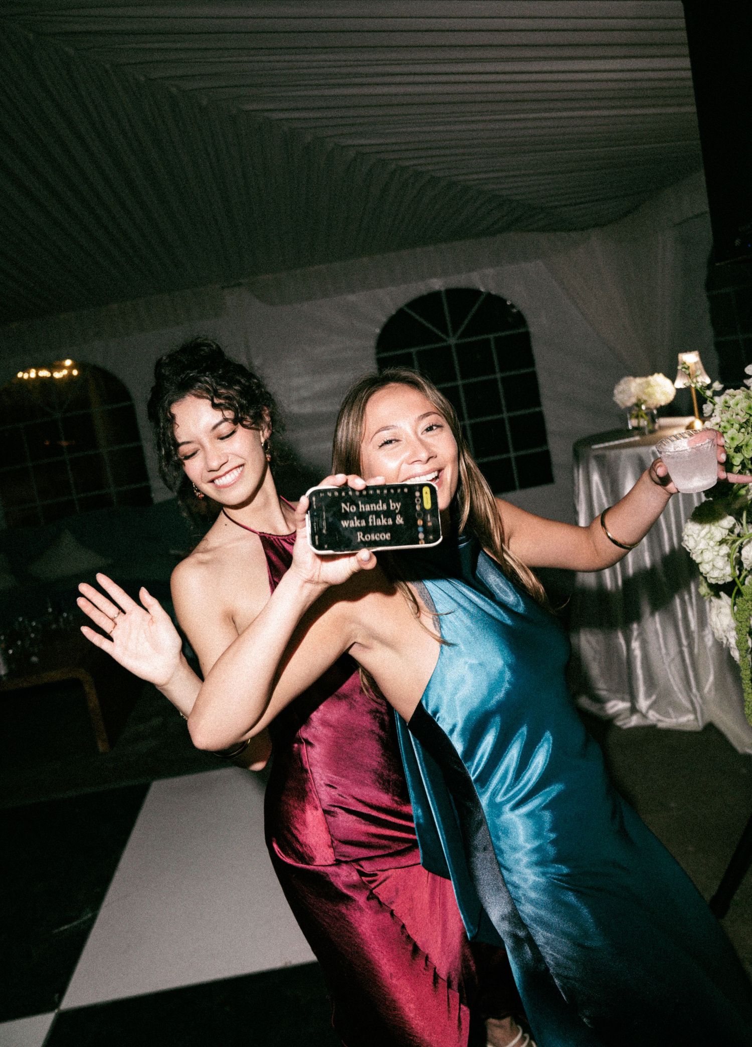 Two guests in colorful gowns posing with phone on wedding dance floor at night.