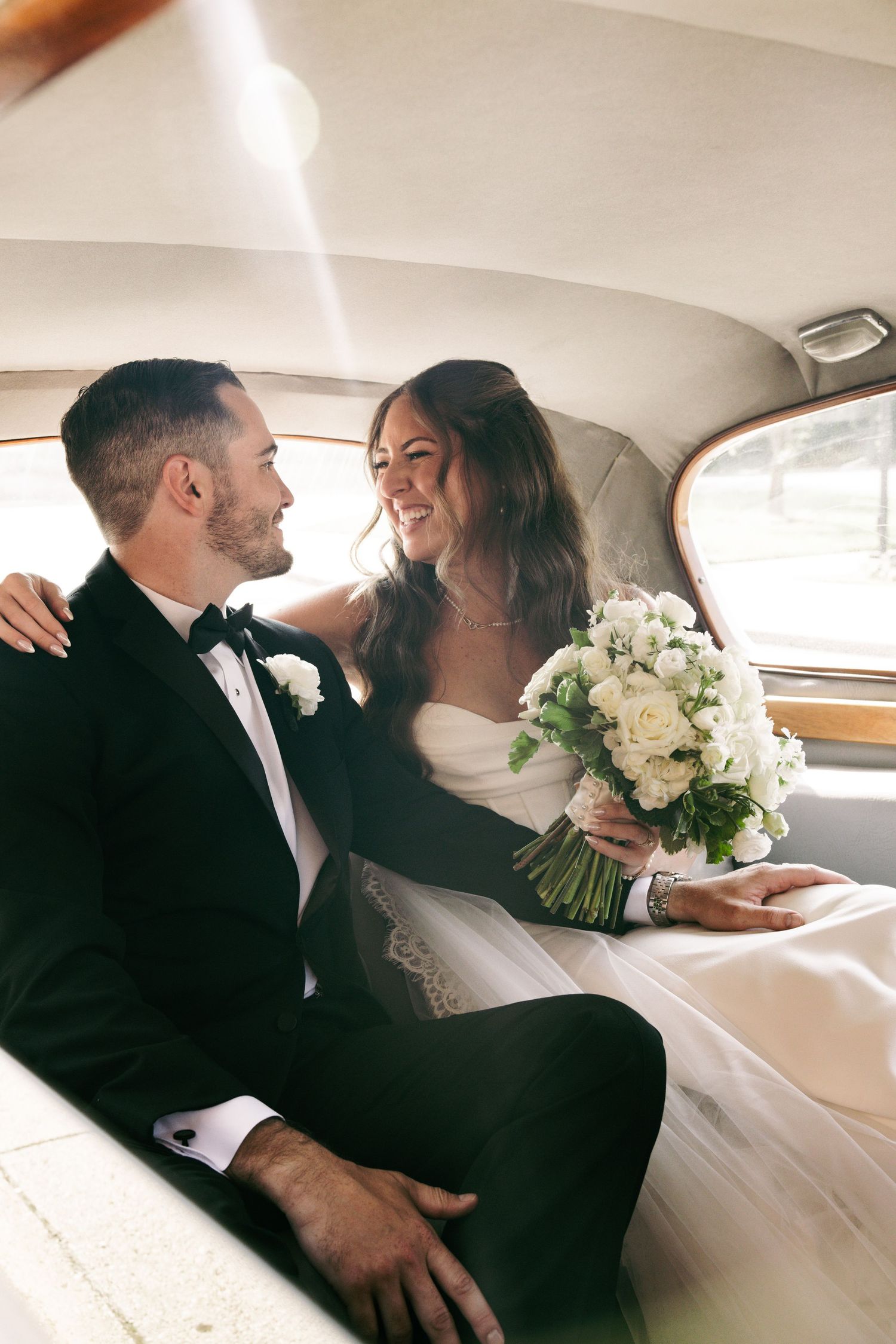 Bride and groom sitting in vintage car backseat, smiling at each other, bride holding white rose bouquet.