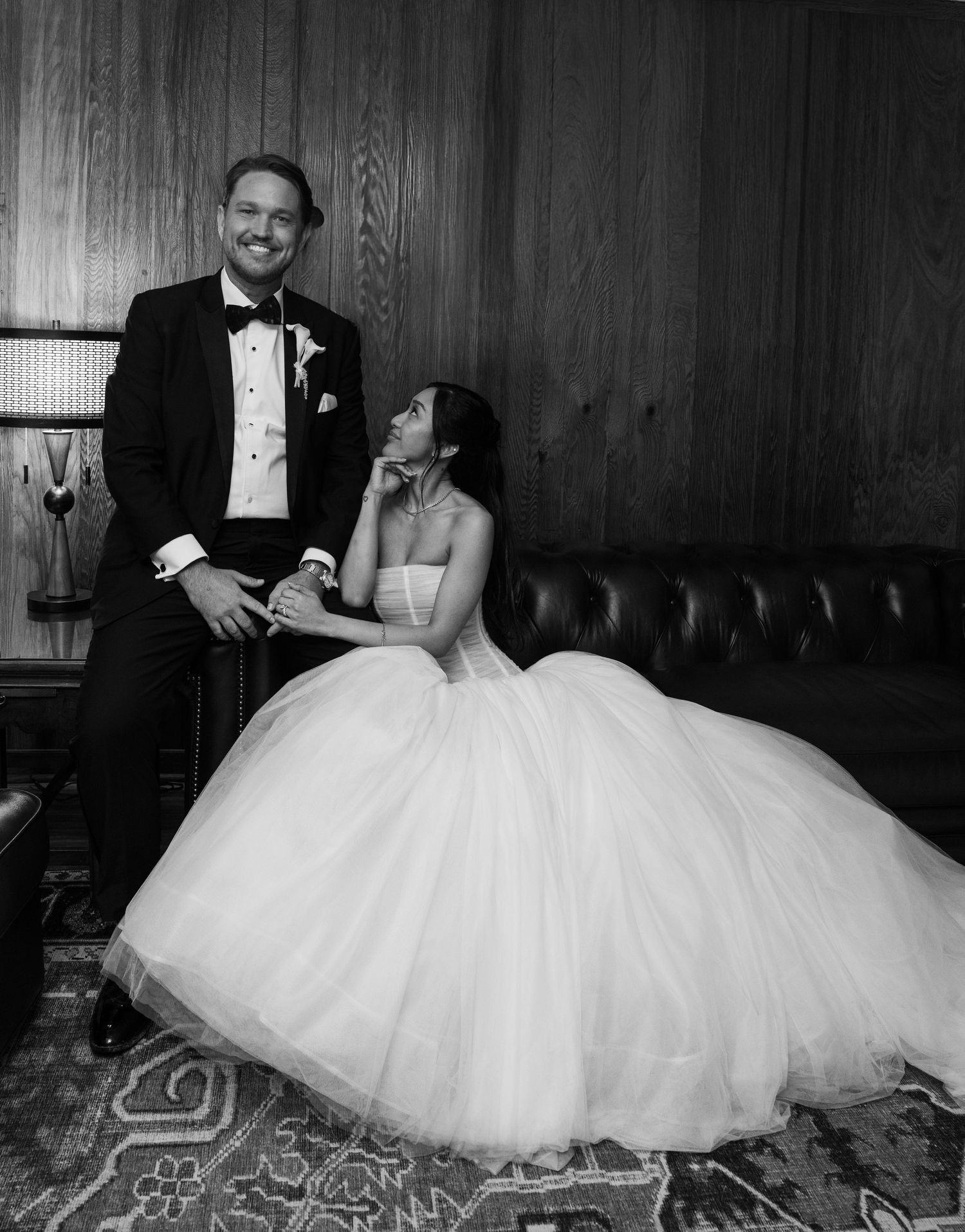 Black and white wedding photo of bride in ballgown seated on leather sofa with groom in tuxedo standing beside her.