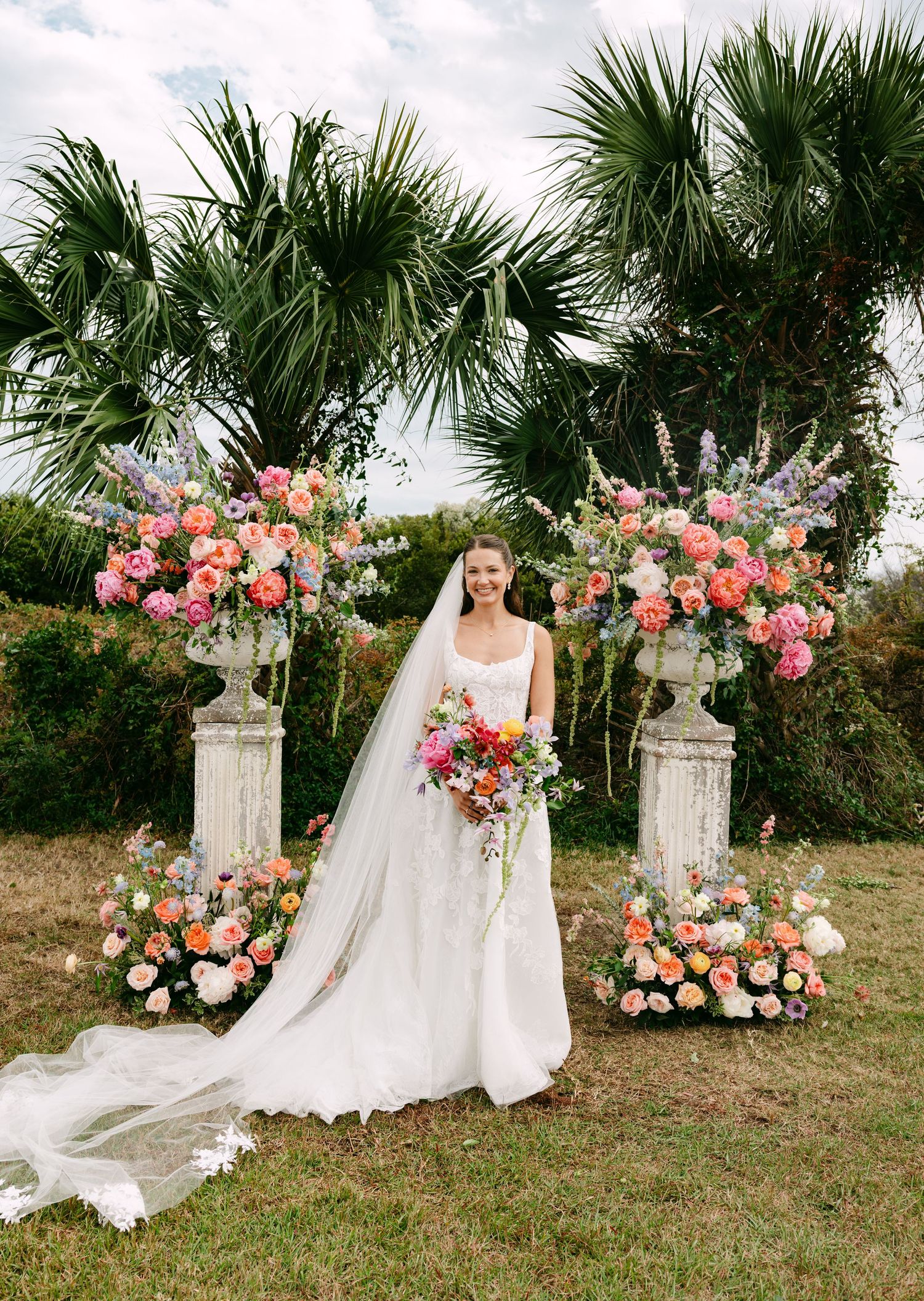 Bride in white gown stands between two ornate floral urns with colorful blooms before tropical palm trees.