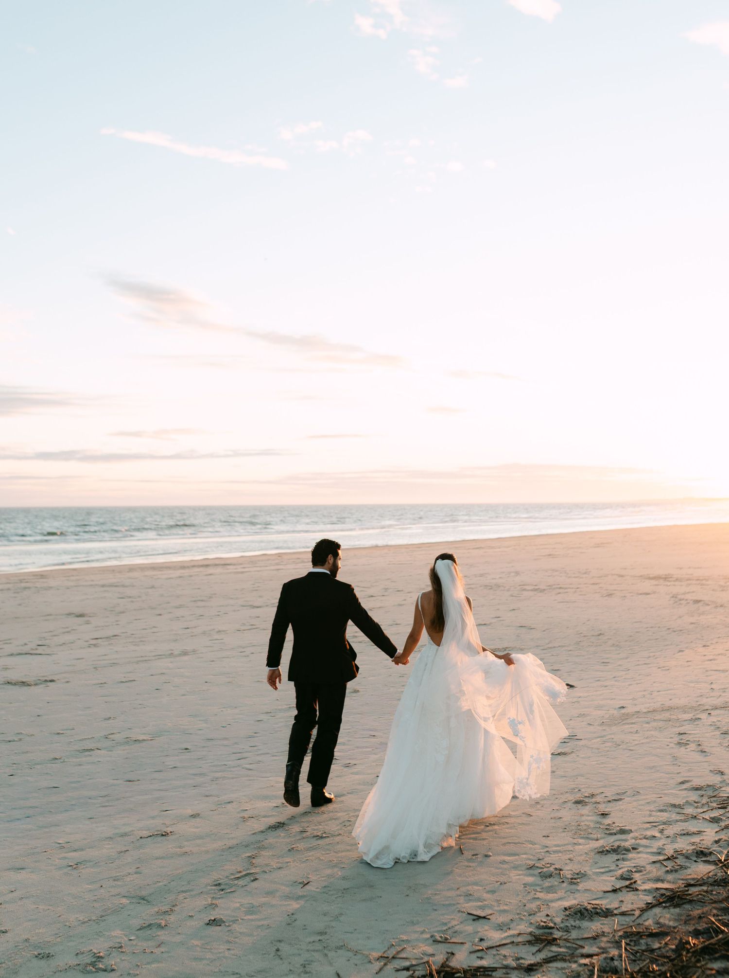 Bride and groom holding hands walking along a sandy beach at sunset in wedding attire with veil flowing in the wind.