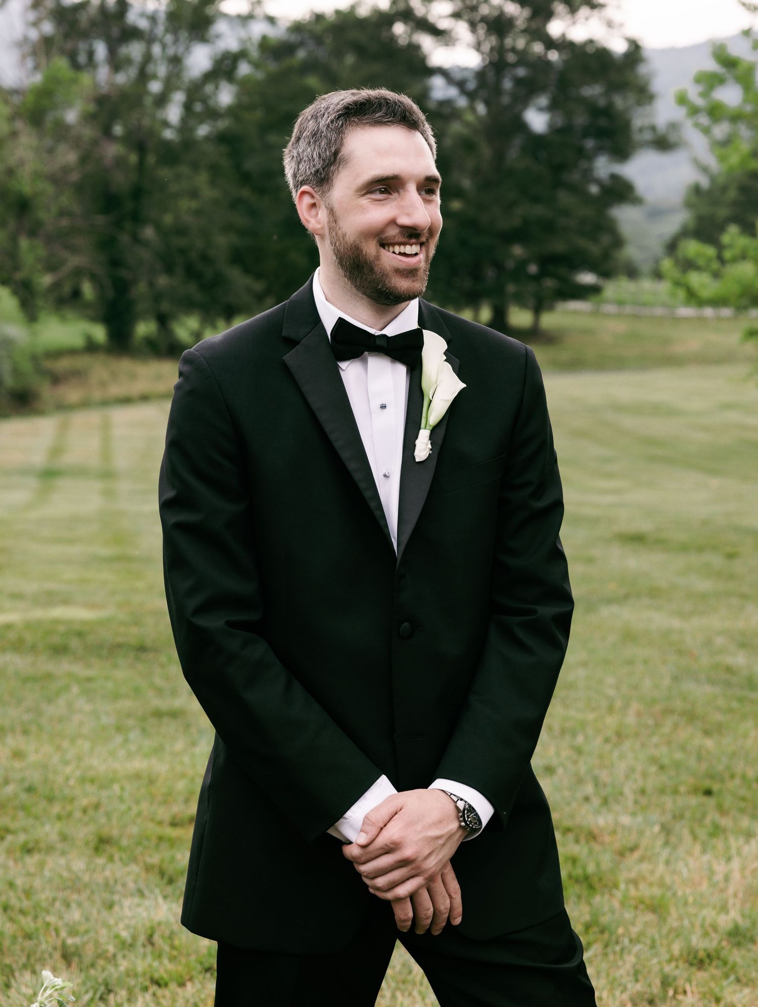 Smiling groom in black tuxedo with bow tie and white boutonniere stands on green lawn outdoors.