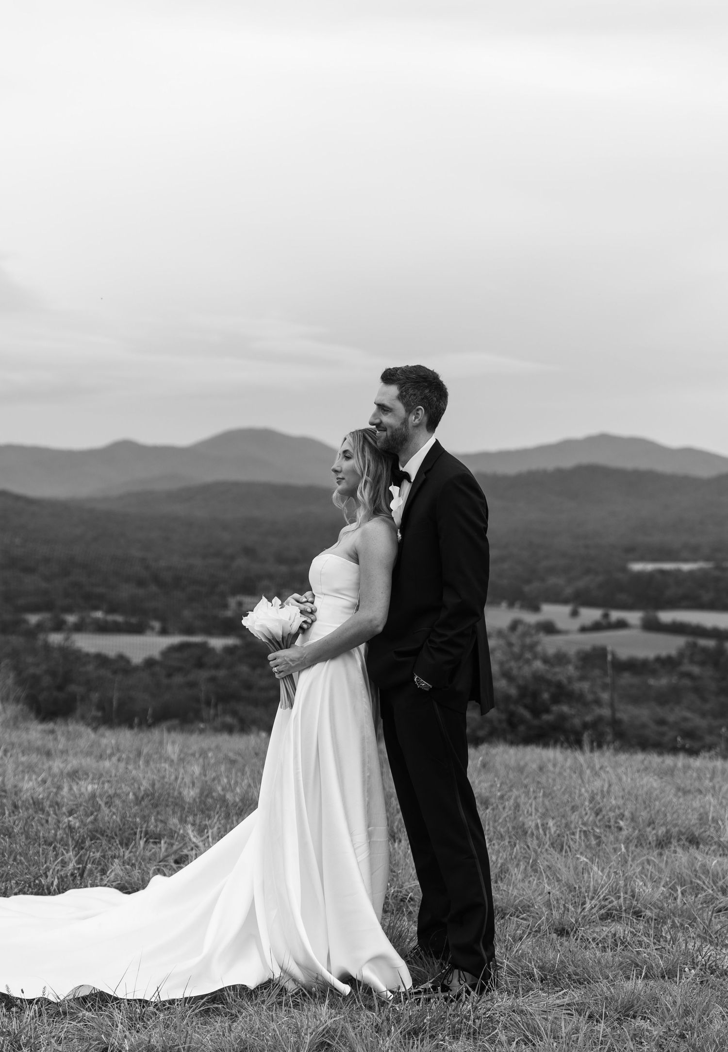 Black and white photo of bride and groom embracing on grassy hilltop with sweeping mountain landscape.
