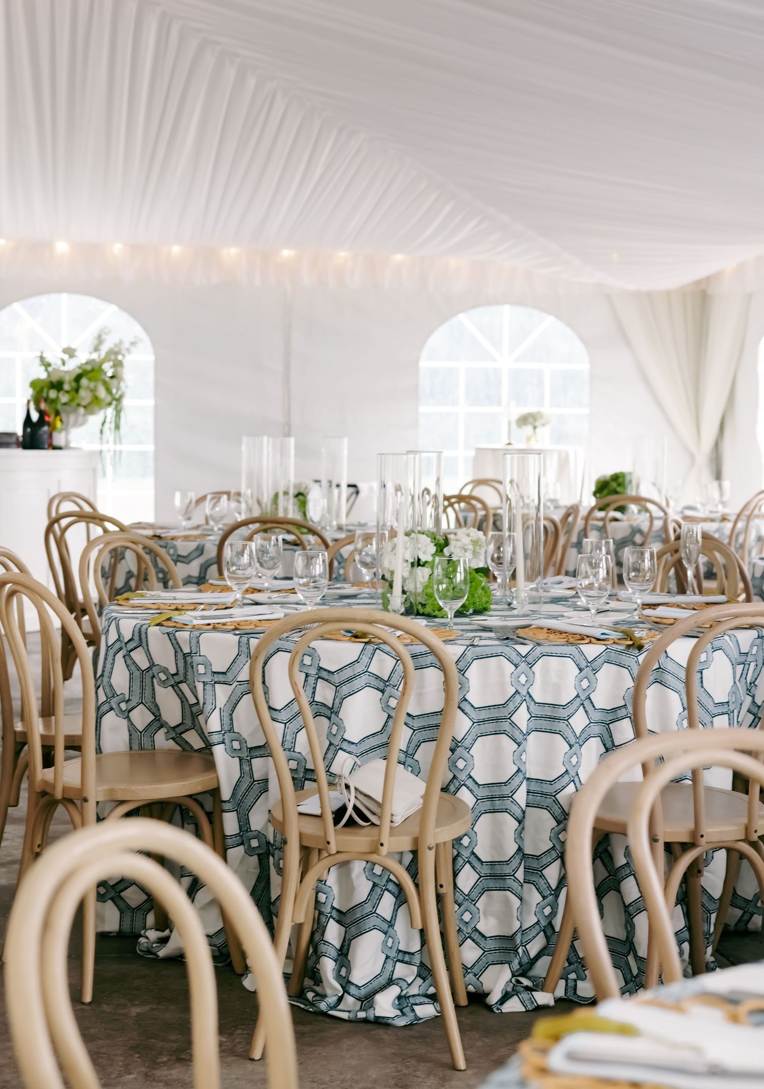 Elegant wedding reception tent with bentwood chairs, patterned tablecloths, crystal glassware, and draped white ceiling with string lights.
