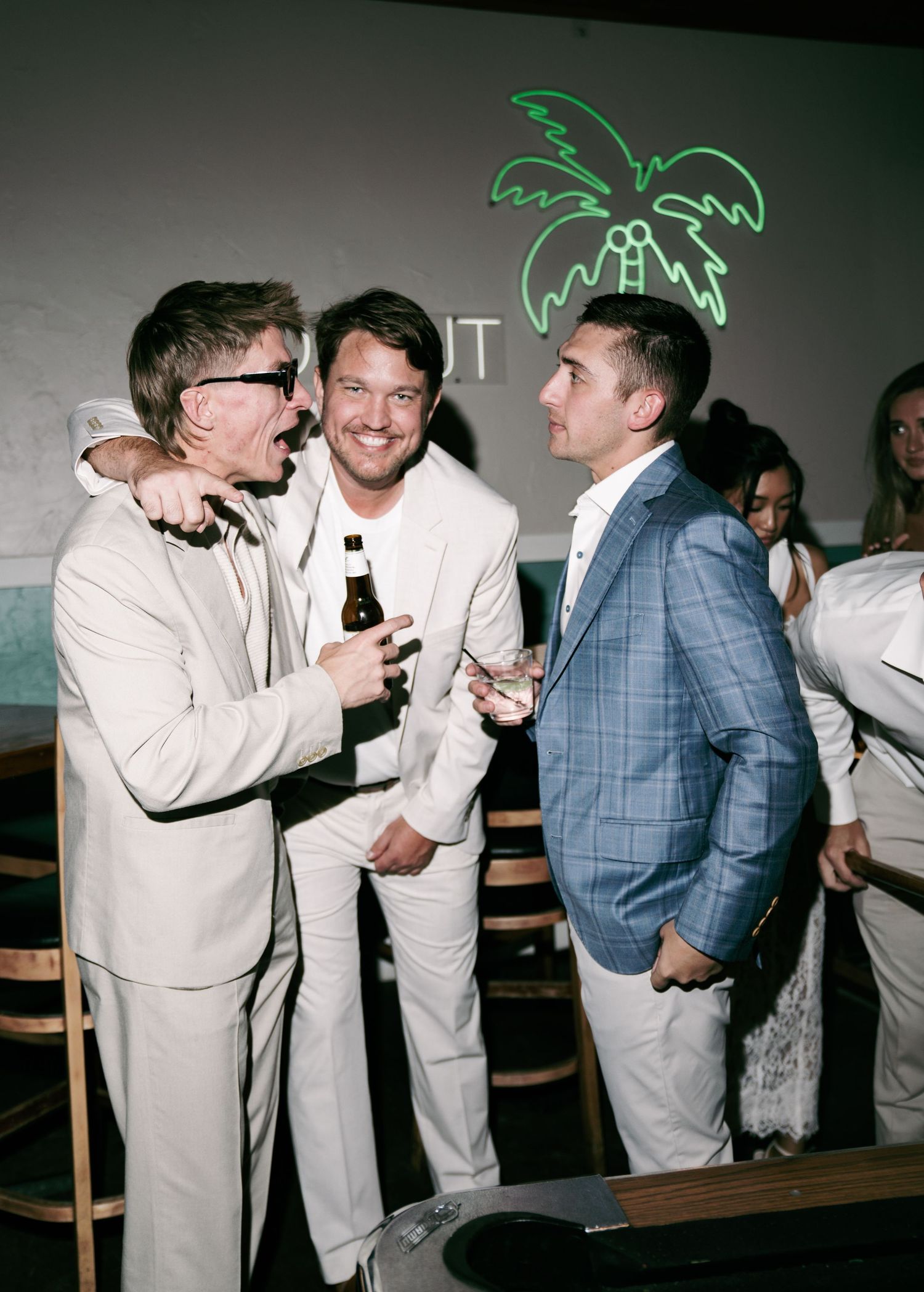 Three men in suits socializing at an upscale event with a neon palm tree sign in the background.
