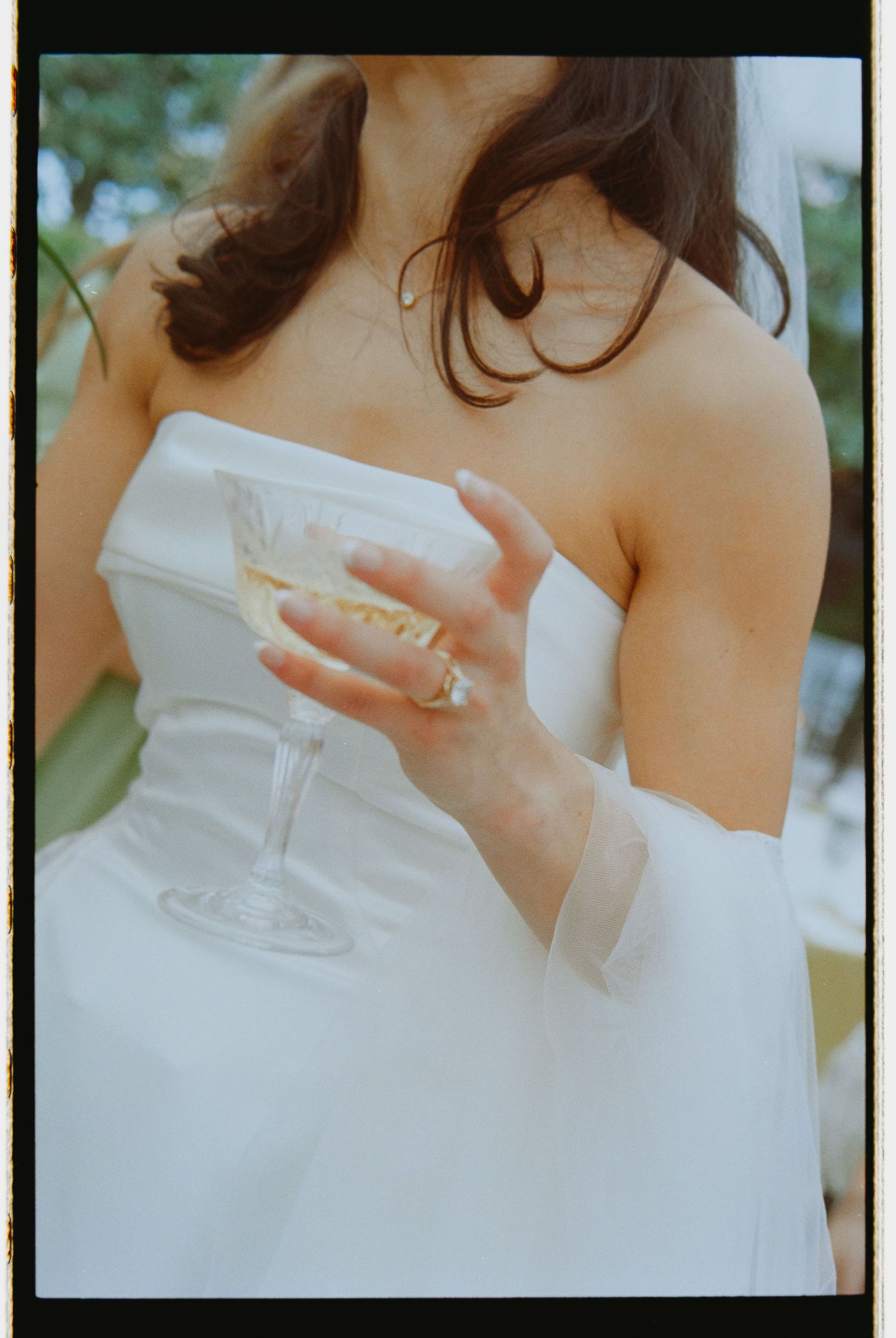 Close-up of bride in strapless white gown holding champagne coupe glass with elegant engagement ring visible.
