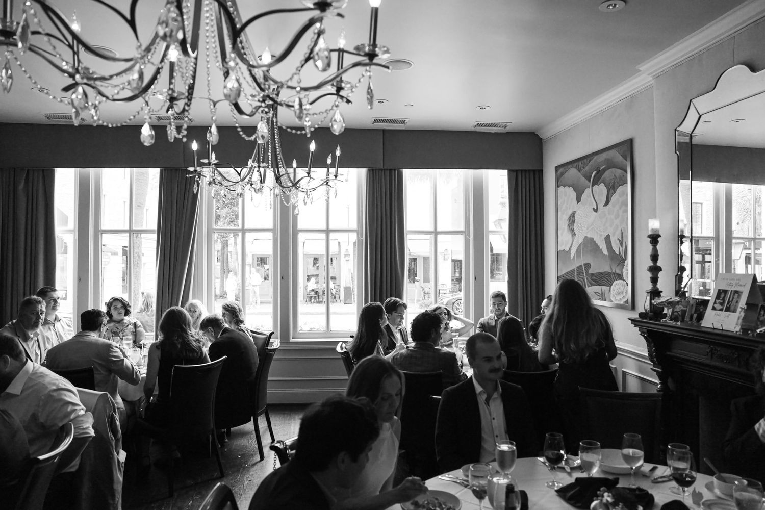 Black and white photo of wedding guests dining in an elegant room with crystal chandelier and large windows.