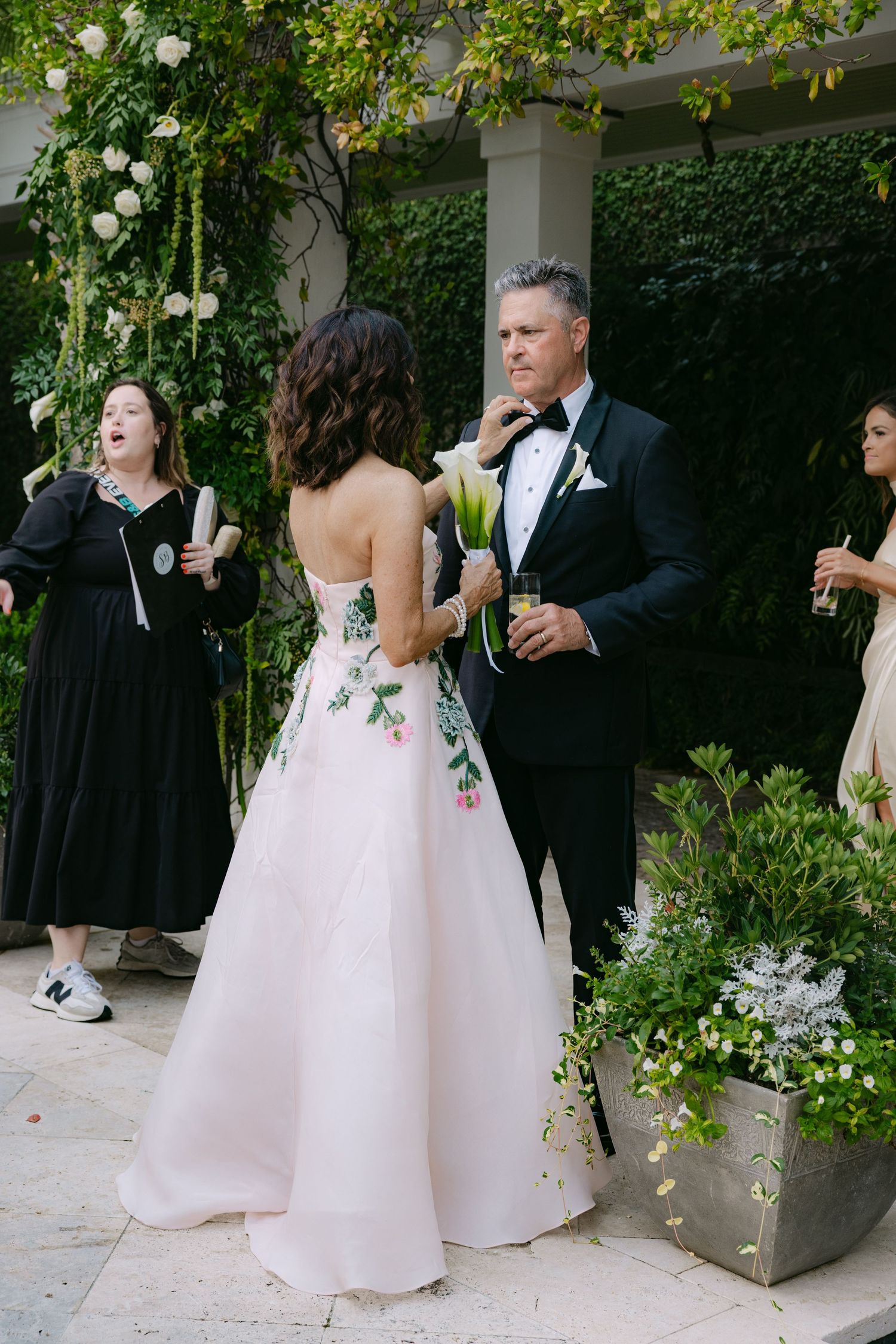 Wedding party members in formal attire socialize outdoors near garden walls with string lights.