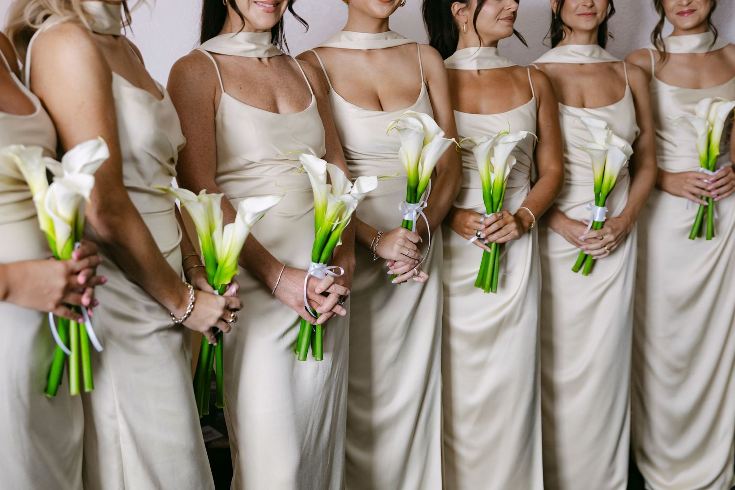 Bridesmaids in cream-colored dresses line up holding green and white floral bouquets.