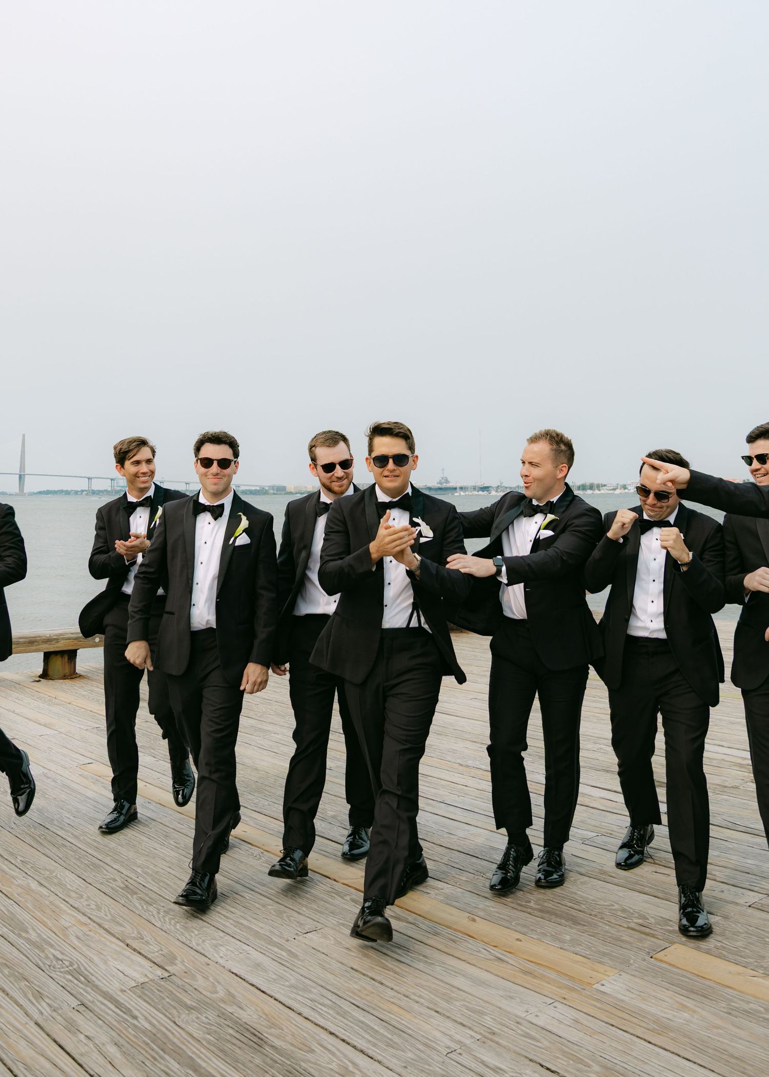 Groomsmen in black tuxedos striking playful poses on wooden boardwalk.