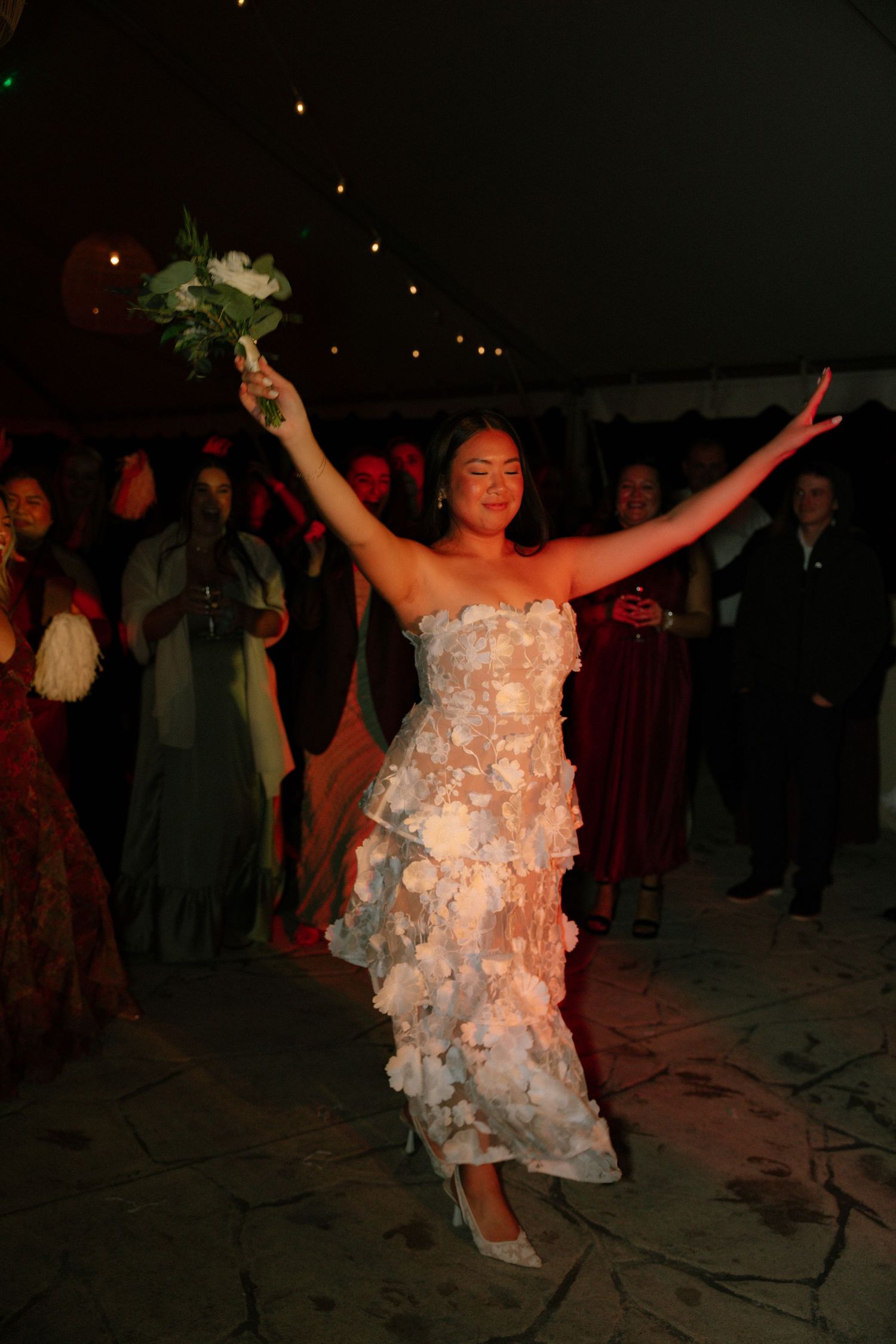 A person in a floral wedding dress dances joyfully at their reception under atmospheric lighting.