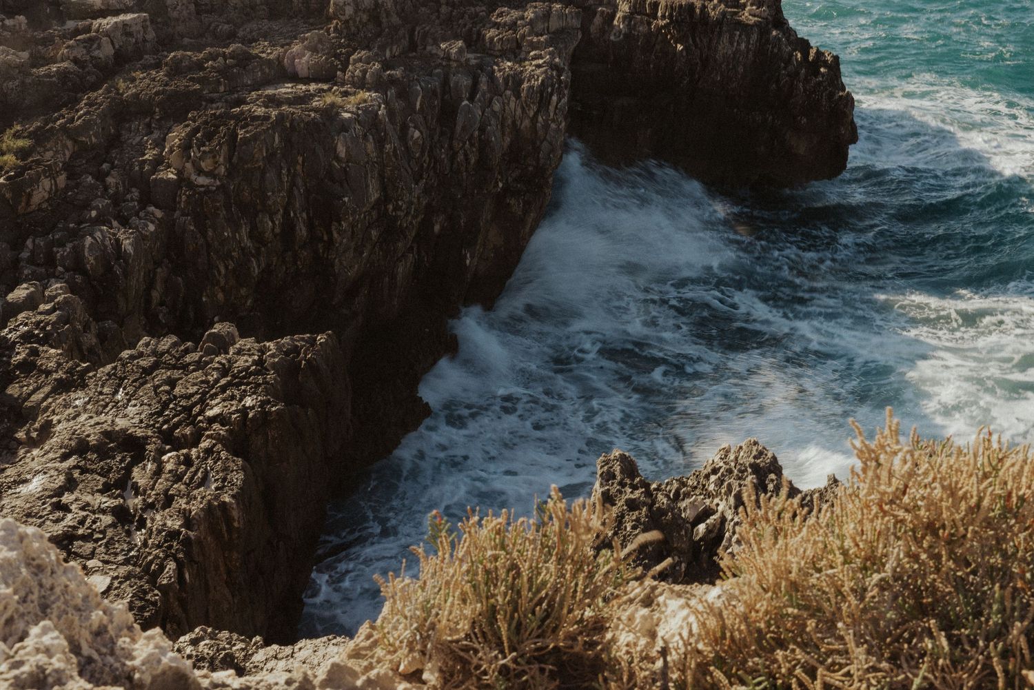 Rocky coastal cliffs tower above turquoise ocean waters as waves crash against the rugged shoreline.