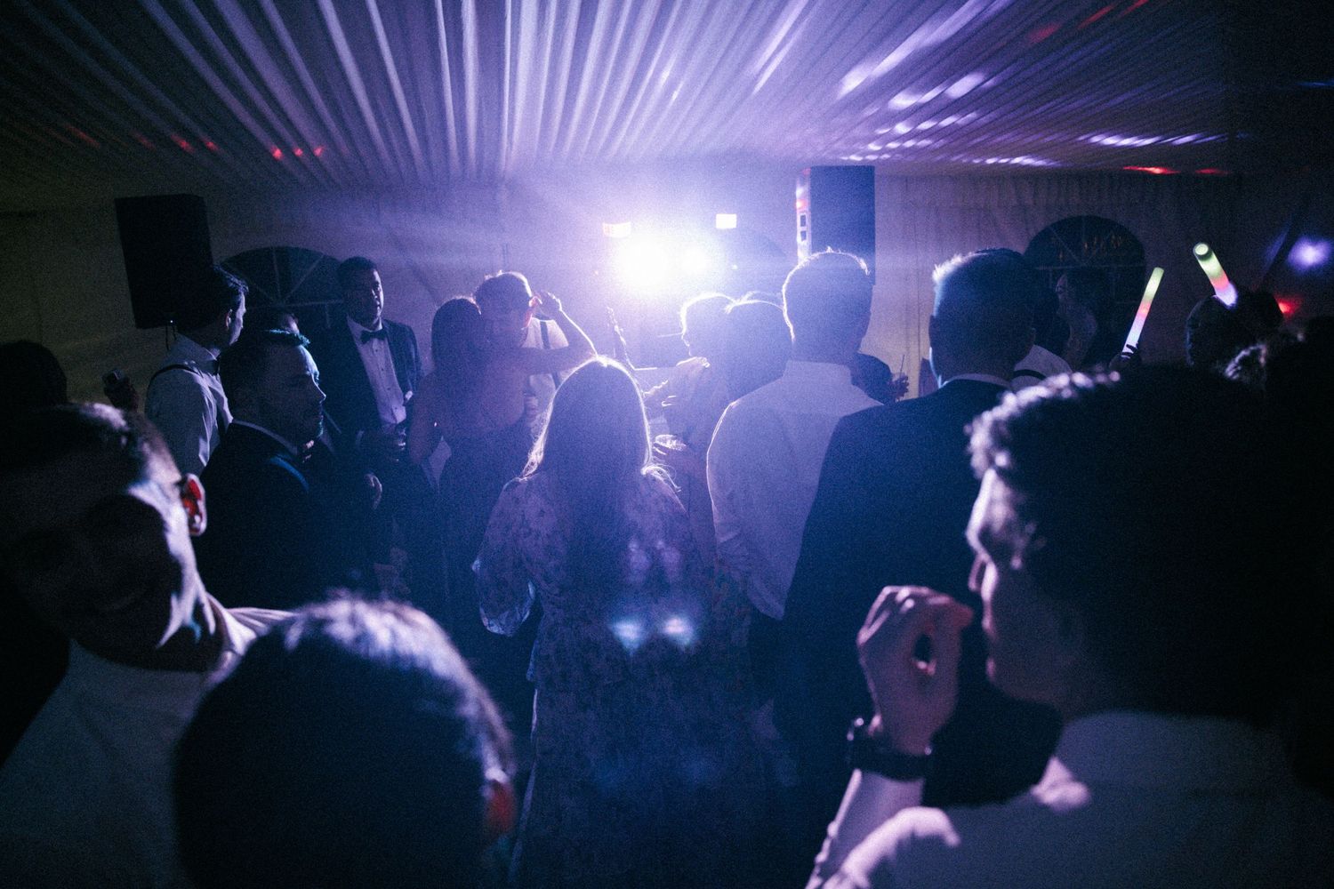 Crowd of people dancing at a dimly lit party with purple stage lighting and bright spotlights in background.
