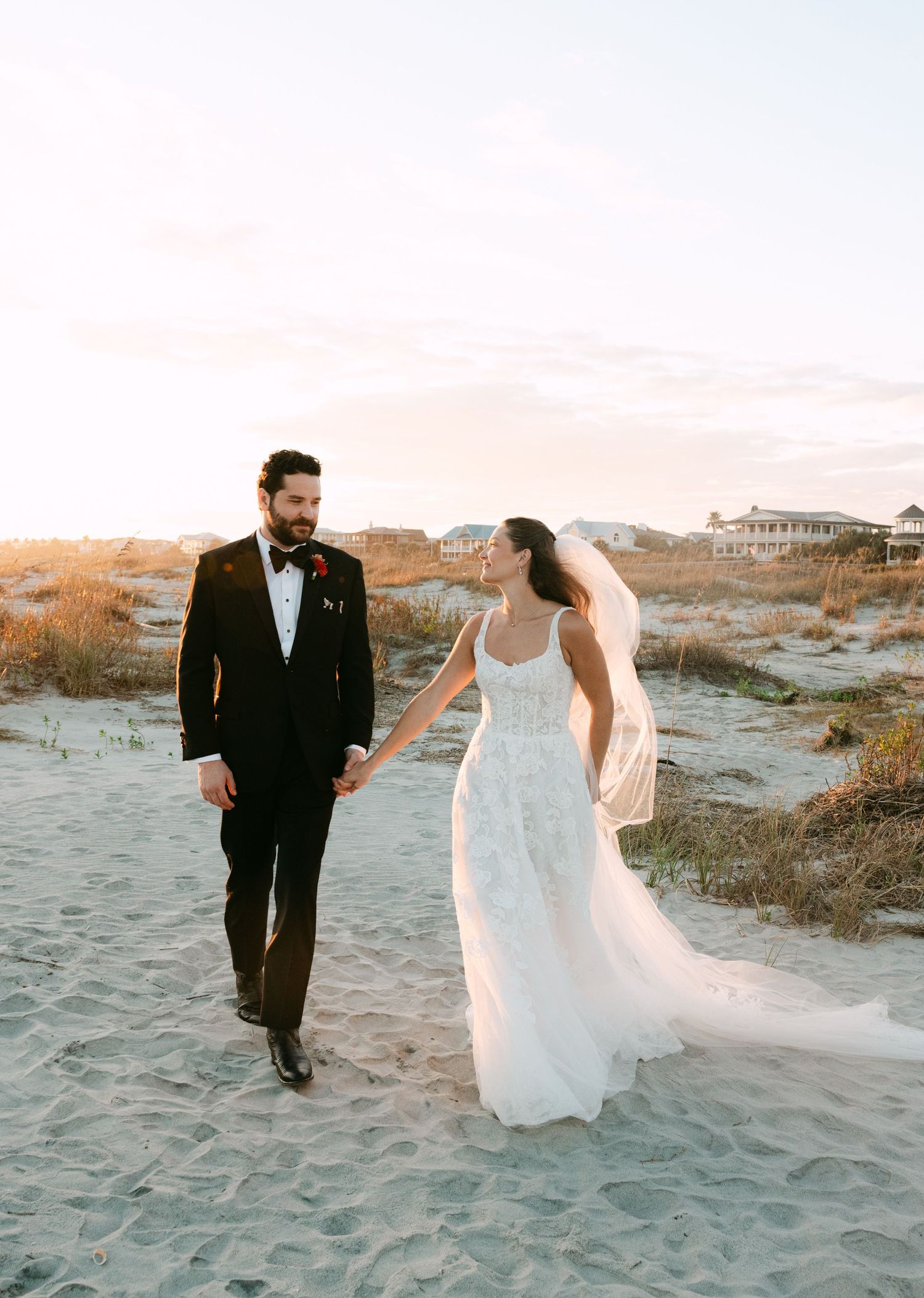 Bride and groom walk hand in hand on sandy beach at golden hour sunset.