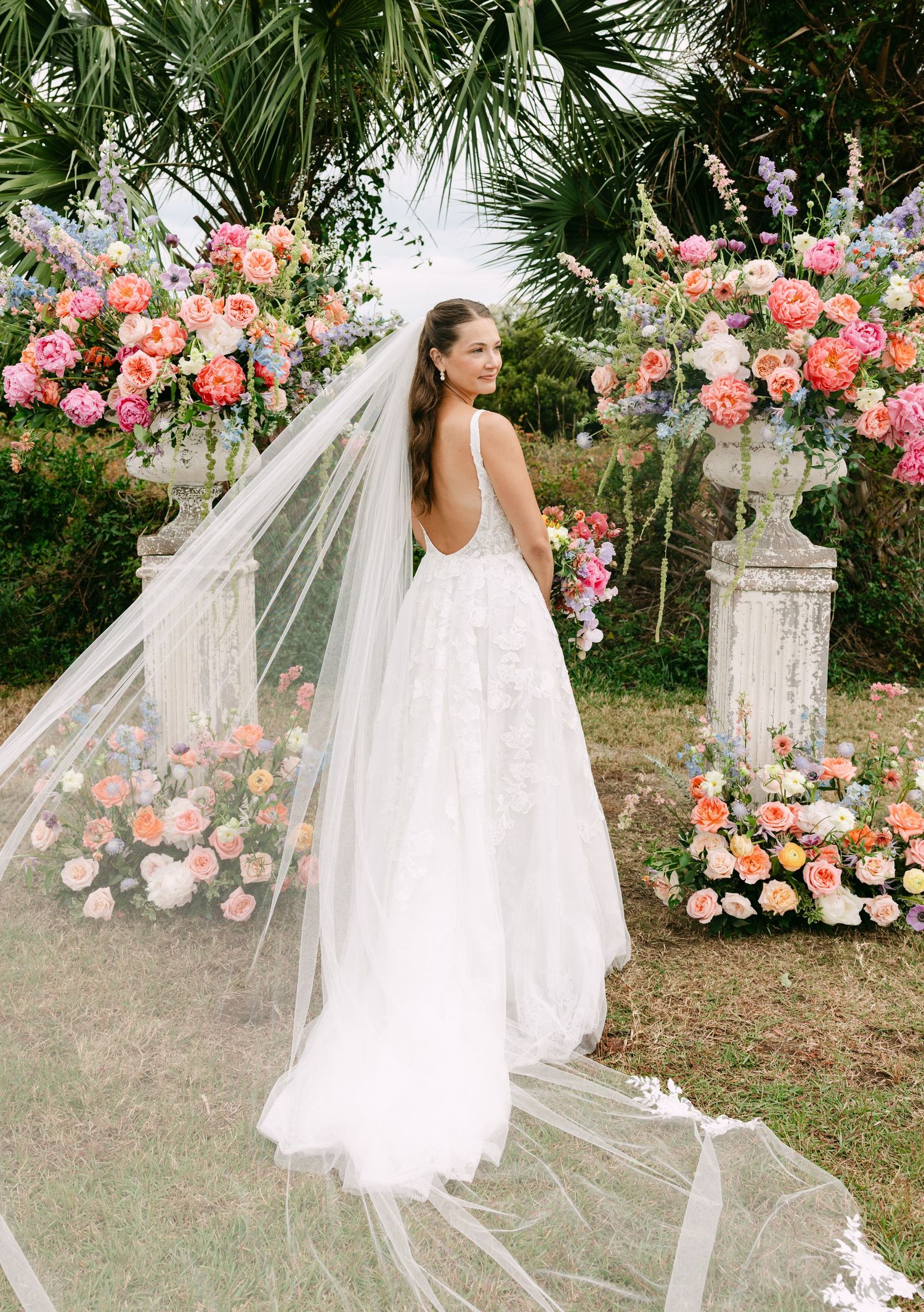 Bride in backless lace wedding gown with long veil stands before colorful floral ceremony arch outdoors.