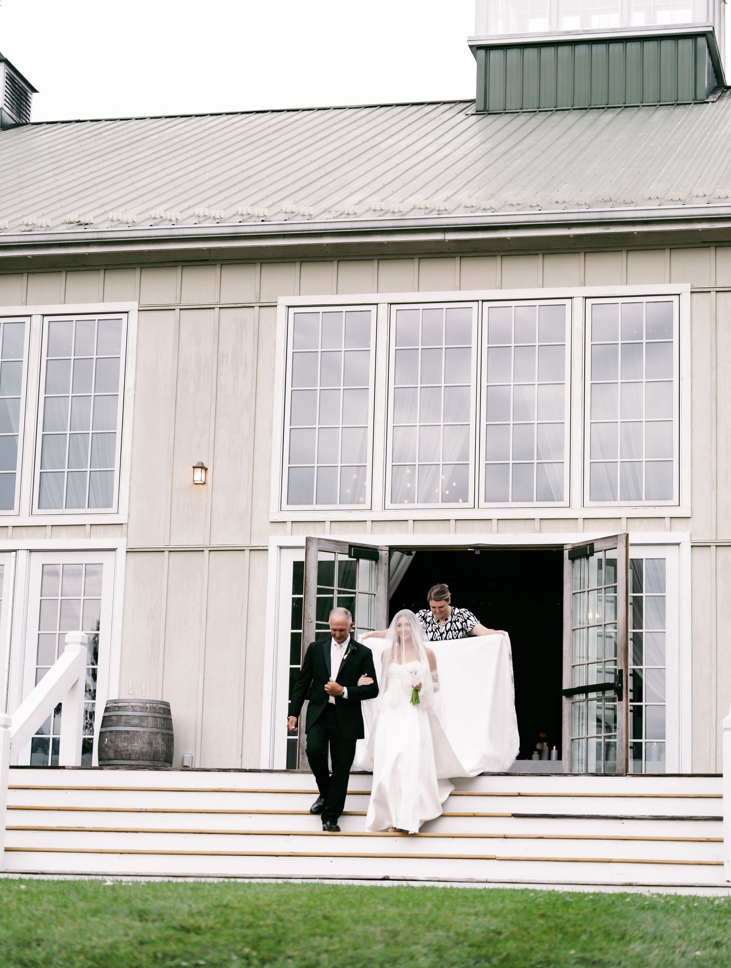 Bride and escort descend steps of rustic barn wedding venue with large windows and green metal roof.