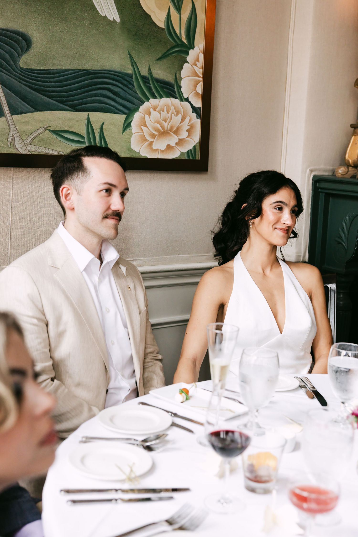 Bride in white halter gown and groom in beige suit seated at wedding dinner table beneath floral wall art.