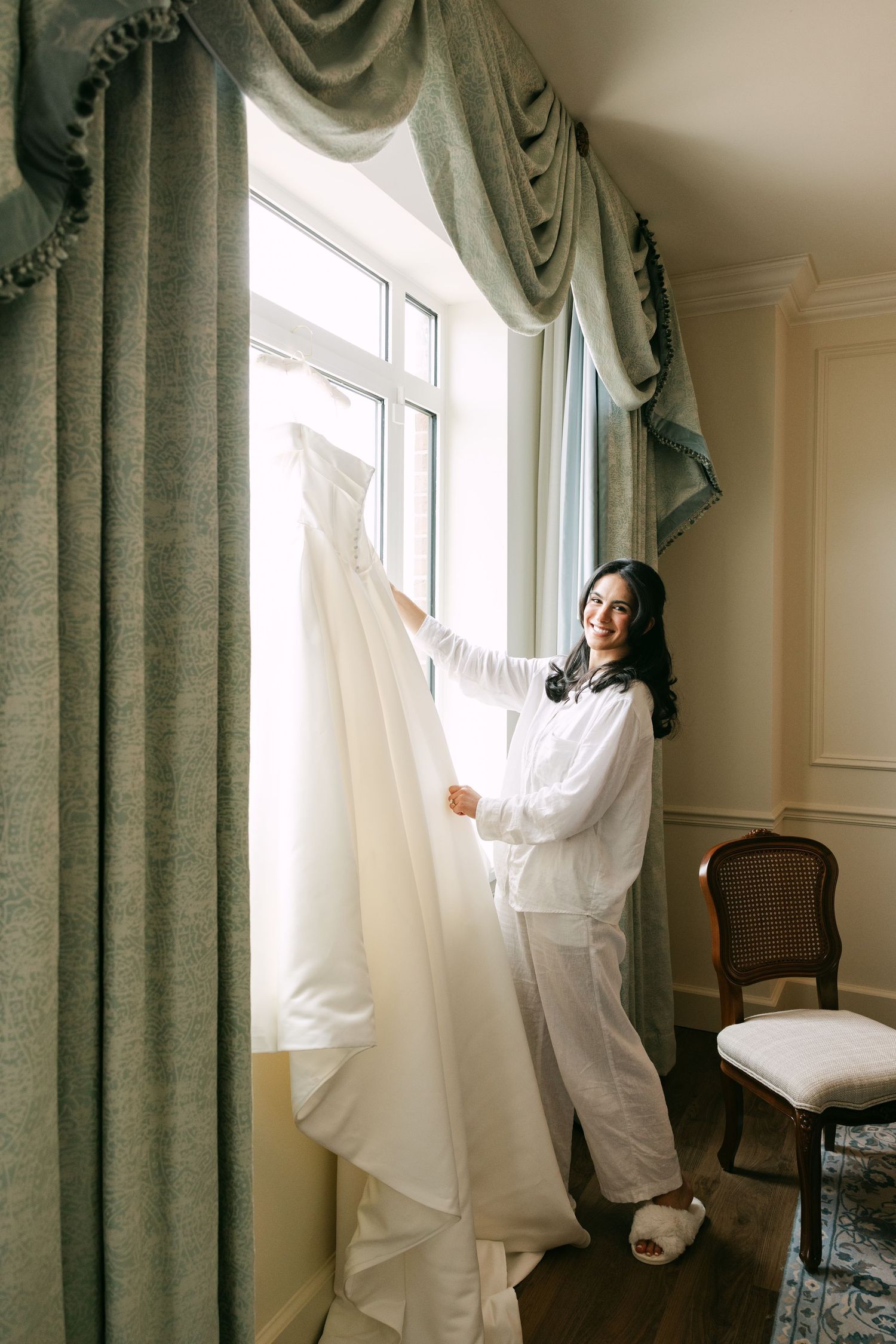 Bride in white robe smiling while holding her wedding dress near an elegant window with green drapes.
