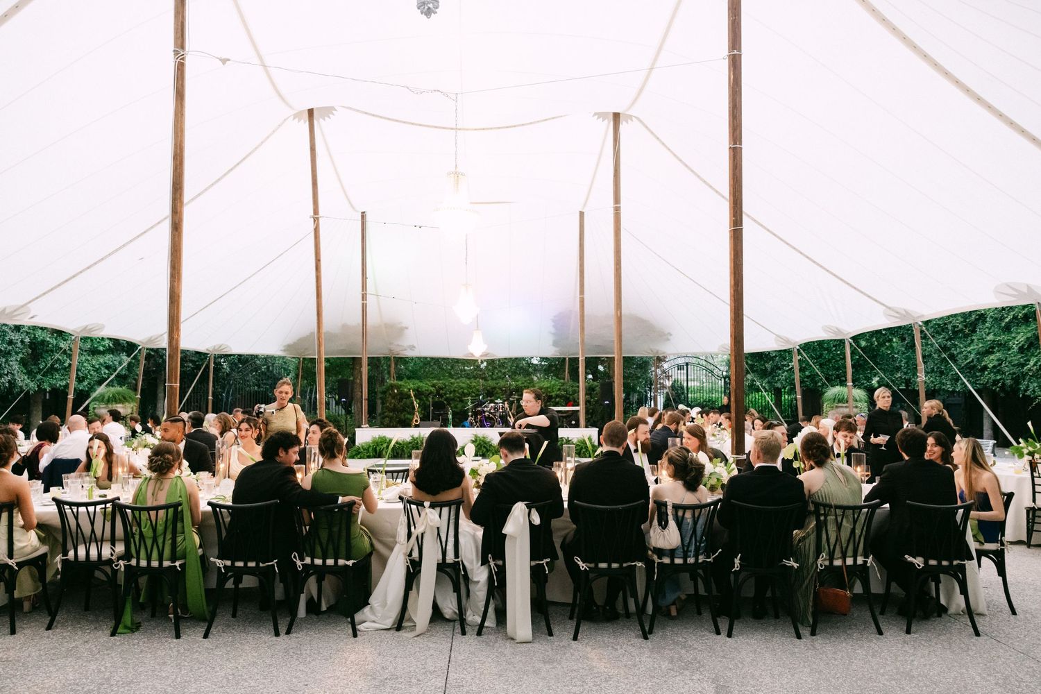 Wedding reception under elegant white sailcloth tent with guests seated at long tables with black chairs.