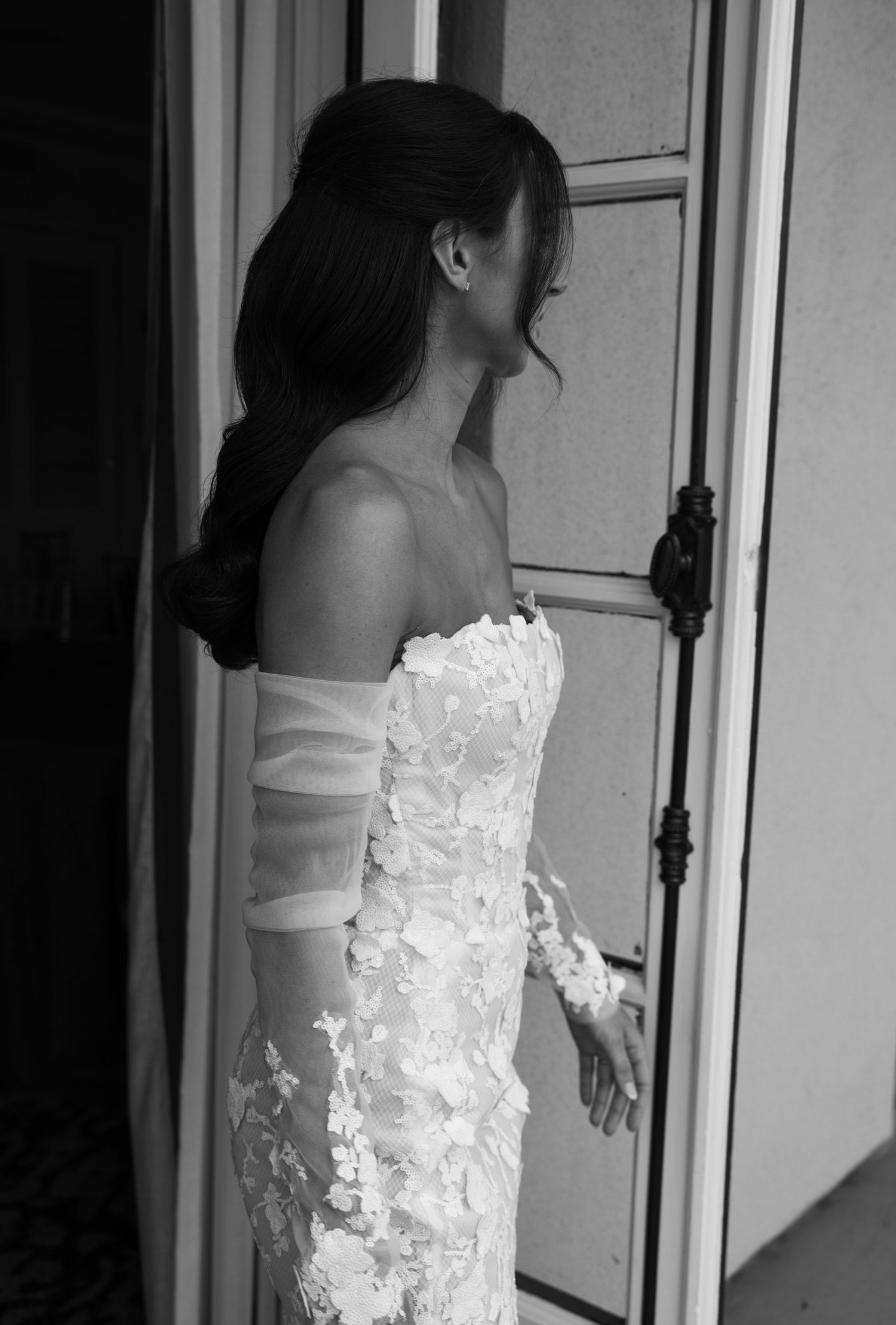A bride in a white lace wedding dress with long sleeves stands by a doorway in black and white.