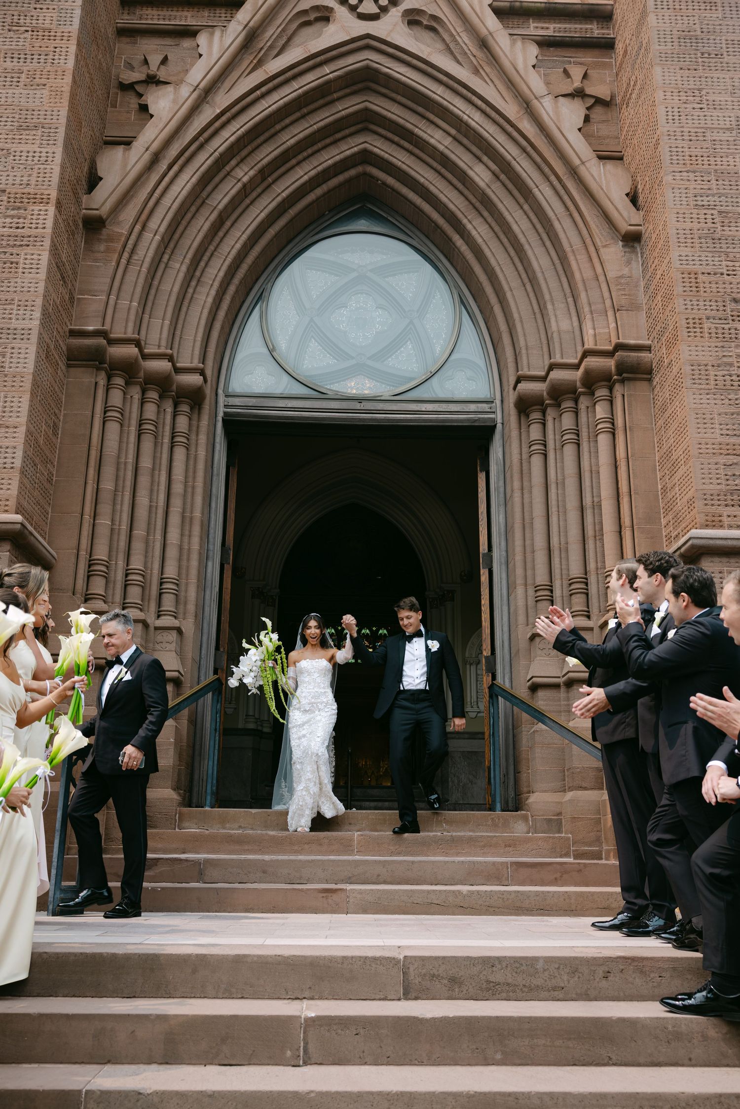 Wedding party descends stone church steps after ceremony while guests look on.