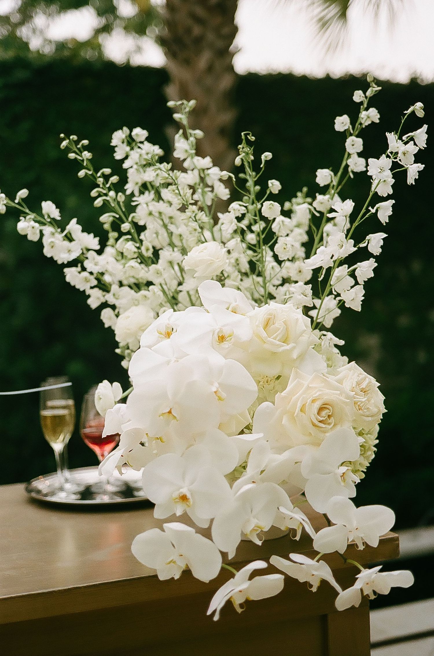 Luxurious white orchid and rose floral arrangements decorate a wooden surface at an outdoor reception.
