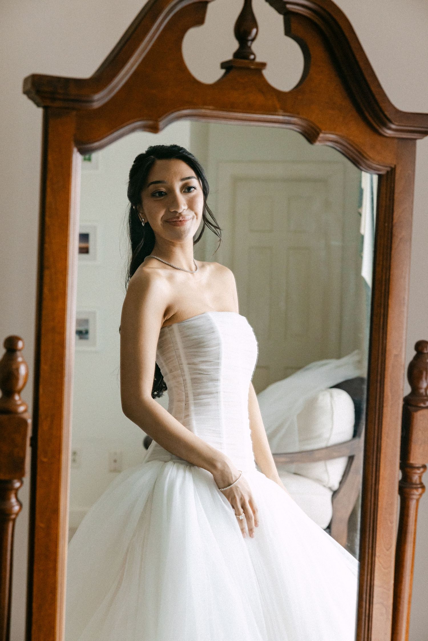 Bride in strapless white ballgown wedding dress smiling at her reflection in a vintage wooden mirror.