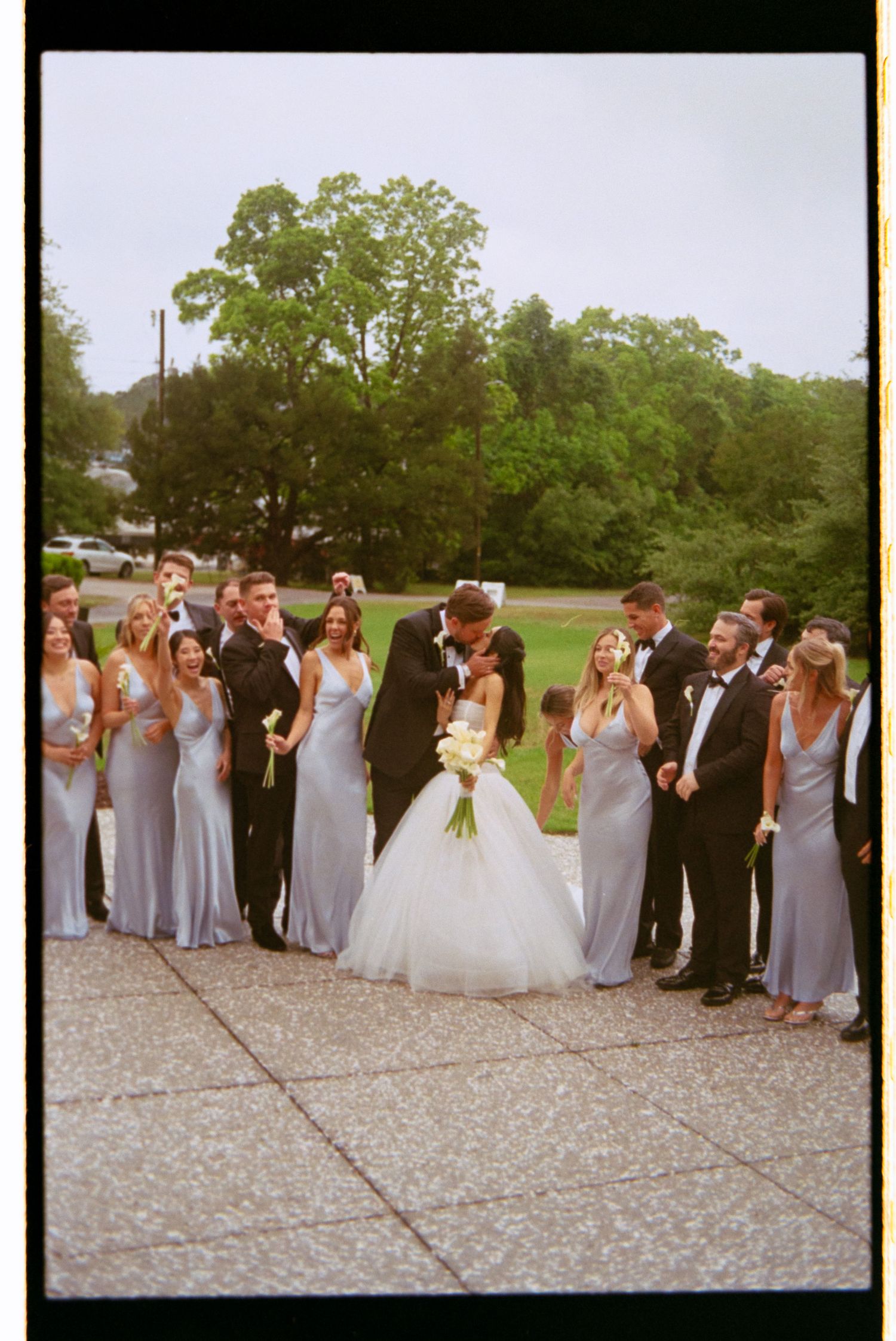 Wedding party poses outdoors as bride and groom kiss, bridesmaids in blue gowns and groomsmen in tuxedos on stone plaza.