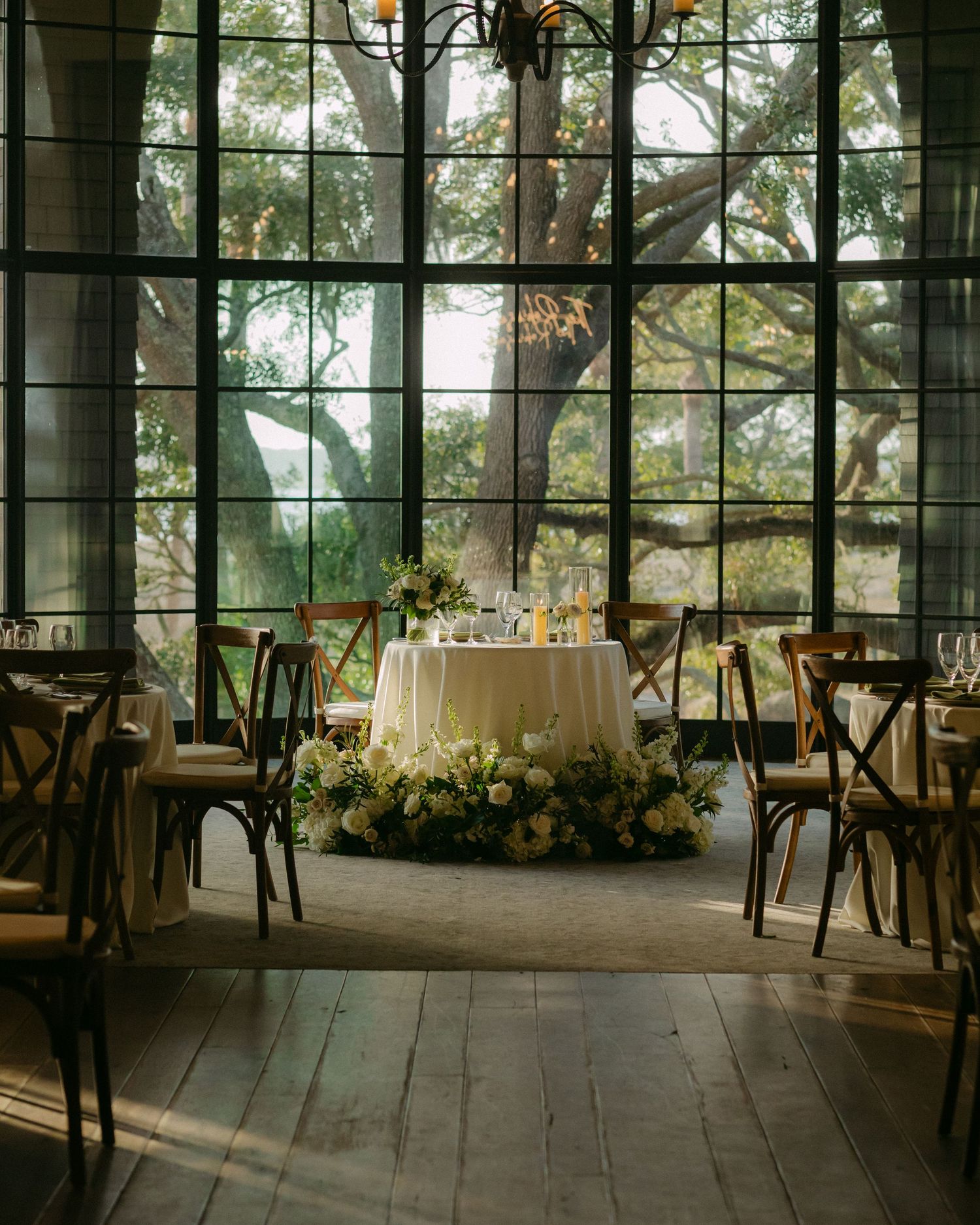 Elegant wedding reception setup with rustic chairs and floral centerpiece in front of large windows.