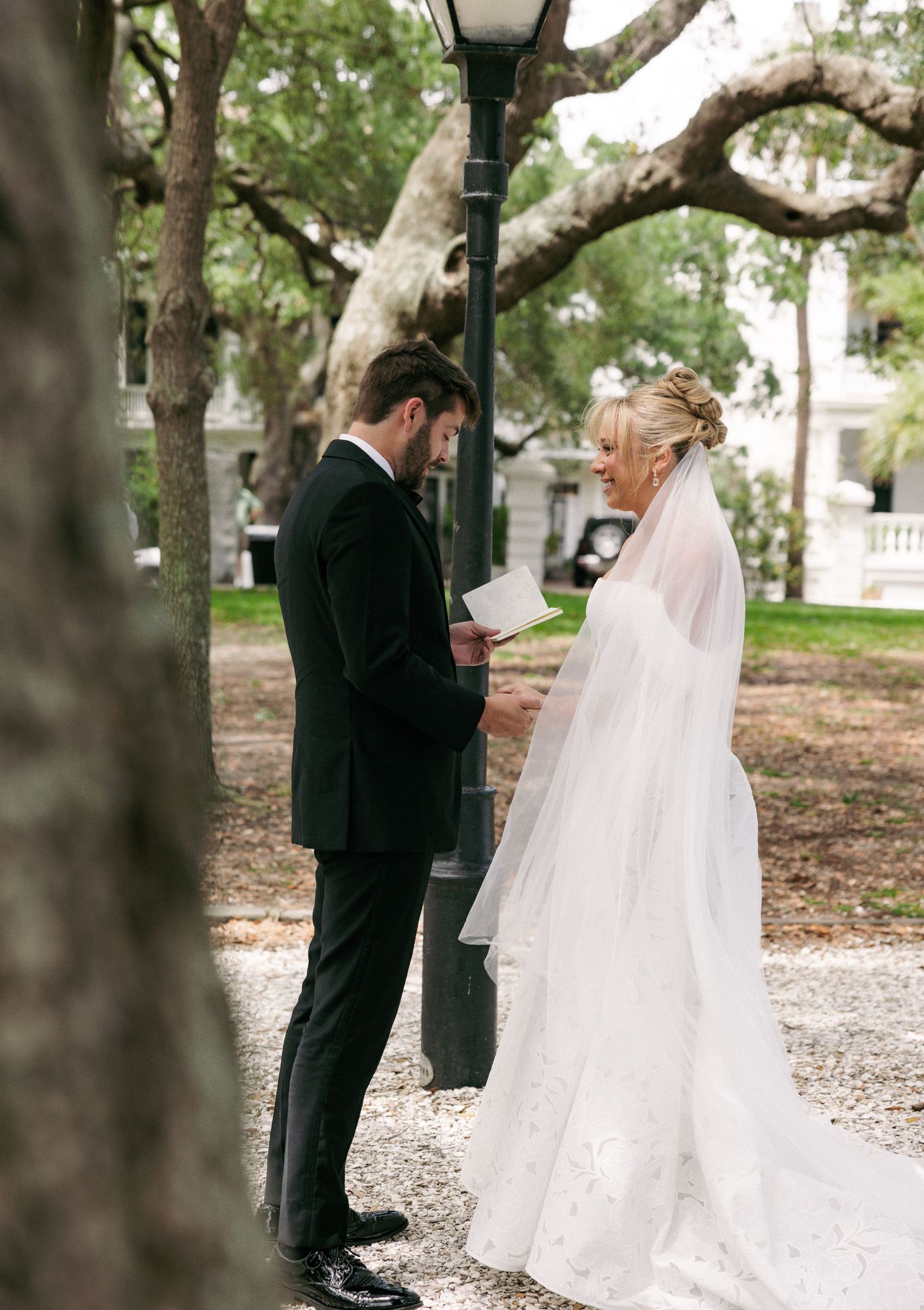 Groom reads wedding vows to bride in white gown and veil outdoors under oak trees near a lamp post.