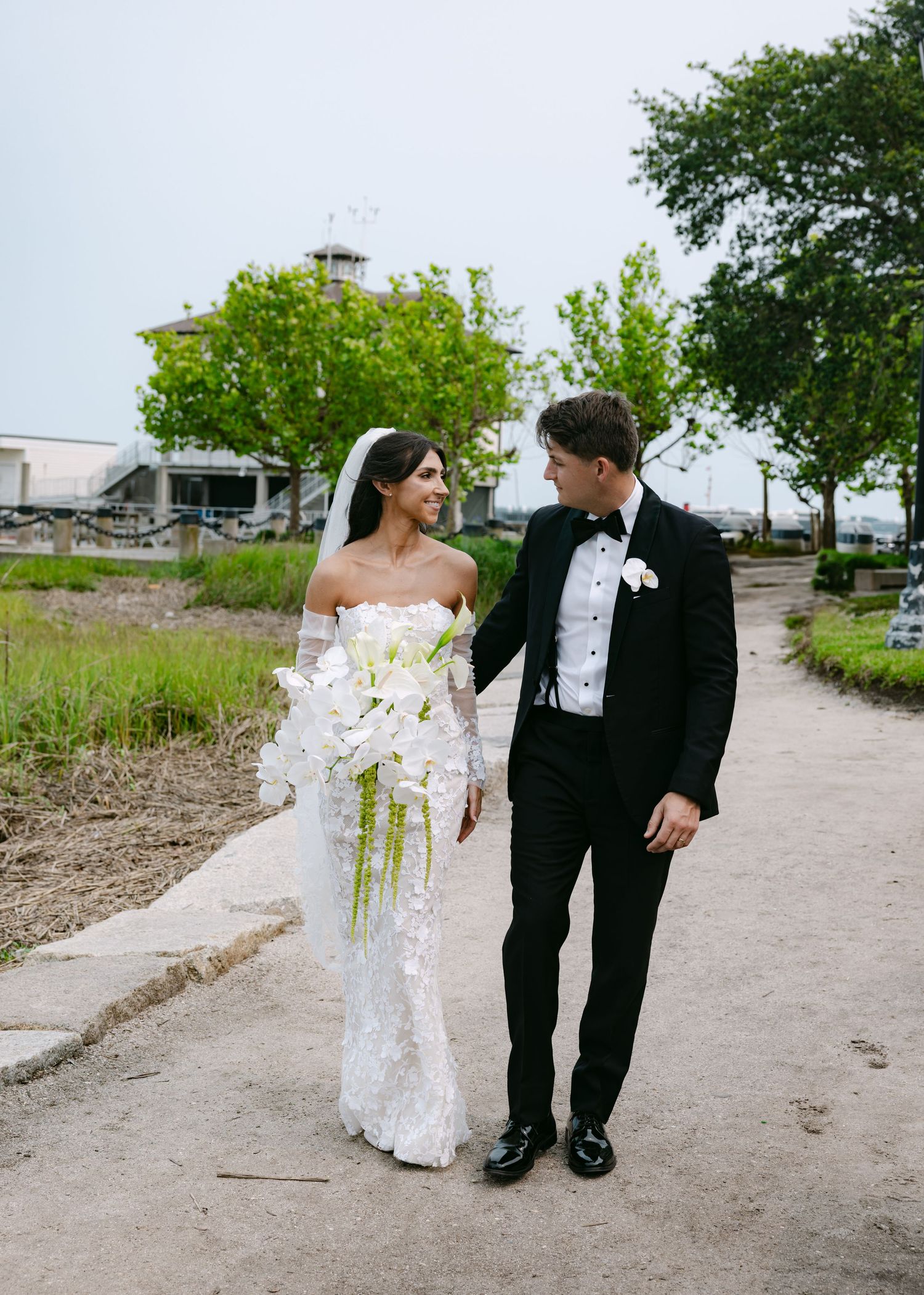 Bride in lace gown and groom in tuxedo walk together on natural pathway during wedding photoshoot.