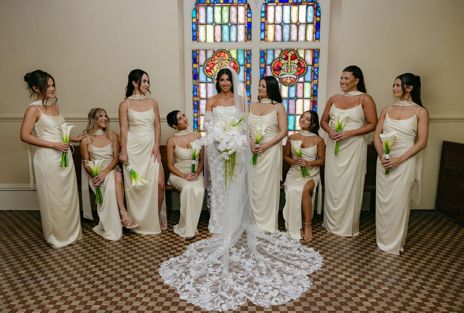 Wedding party lined up in cream-colored dresses against stained glass windows.