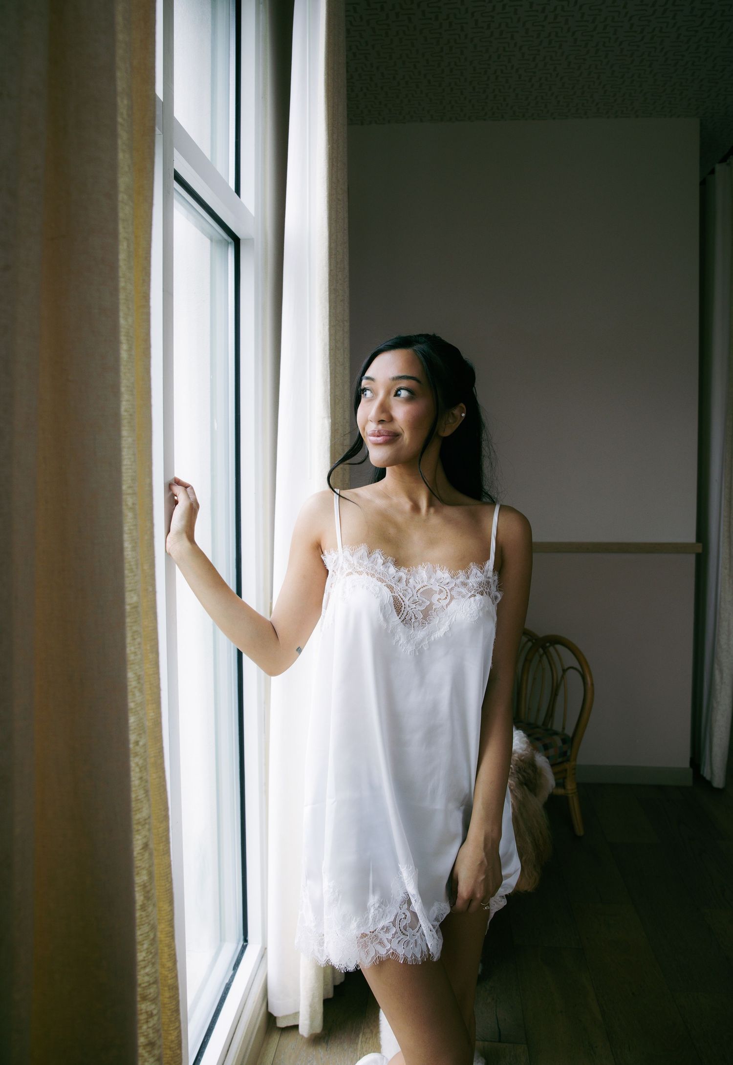 A young woman in a white lace slip dress stands by a sunlit window, smiling softly in a warmly lit room.