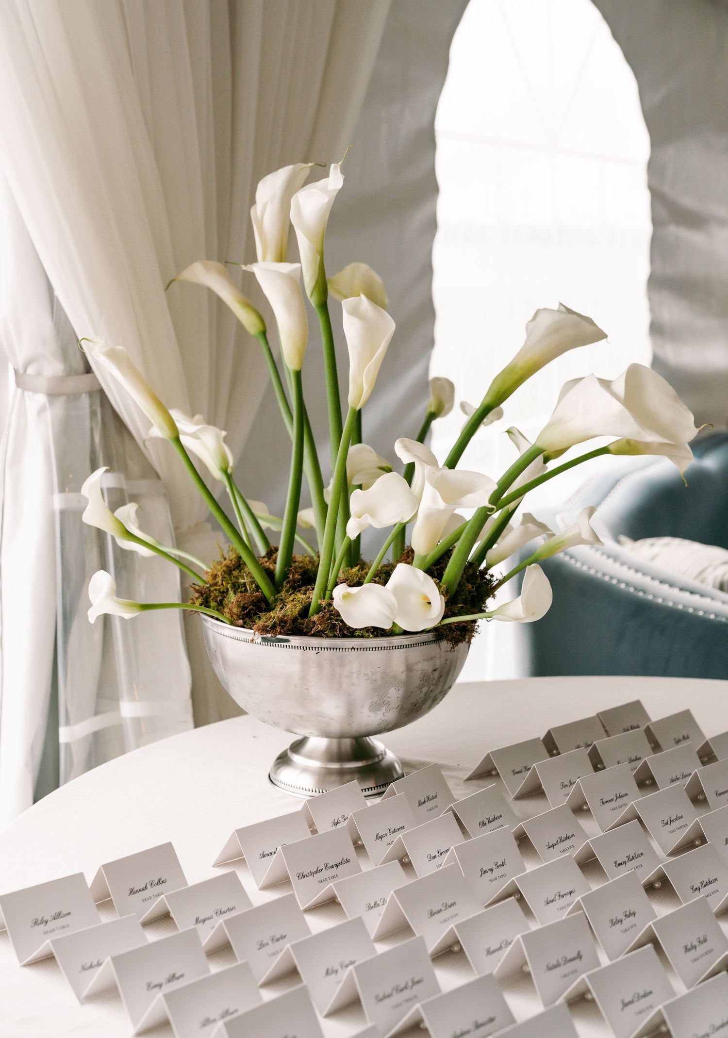 Silver compote vase filled with white calla lilies and moss displayed beside rows of wedding escort cards.