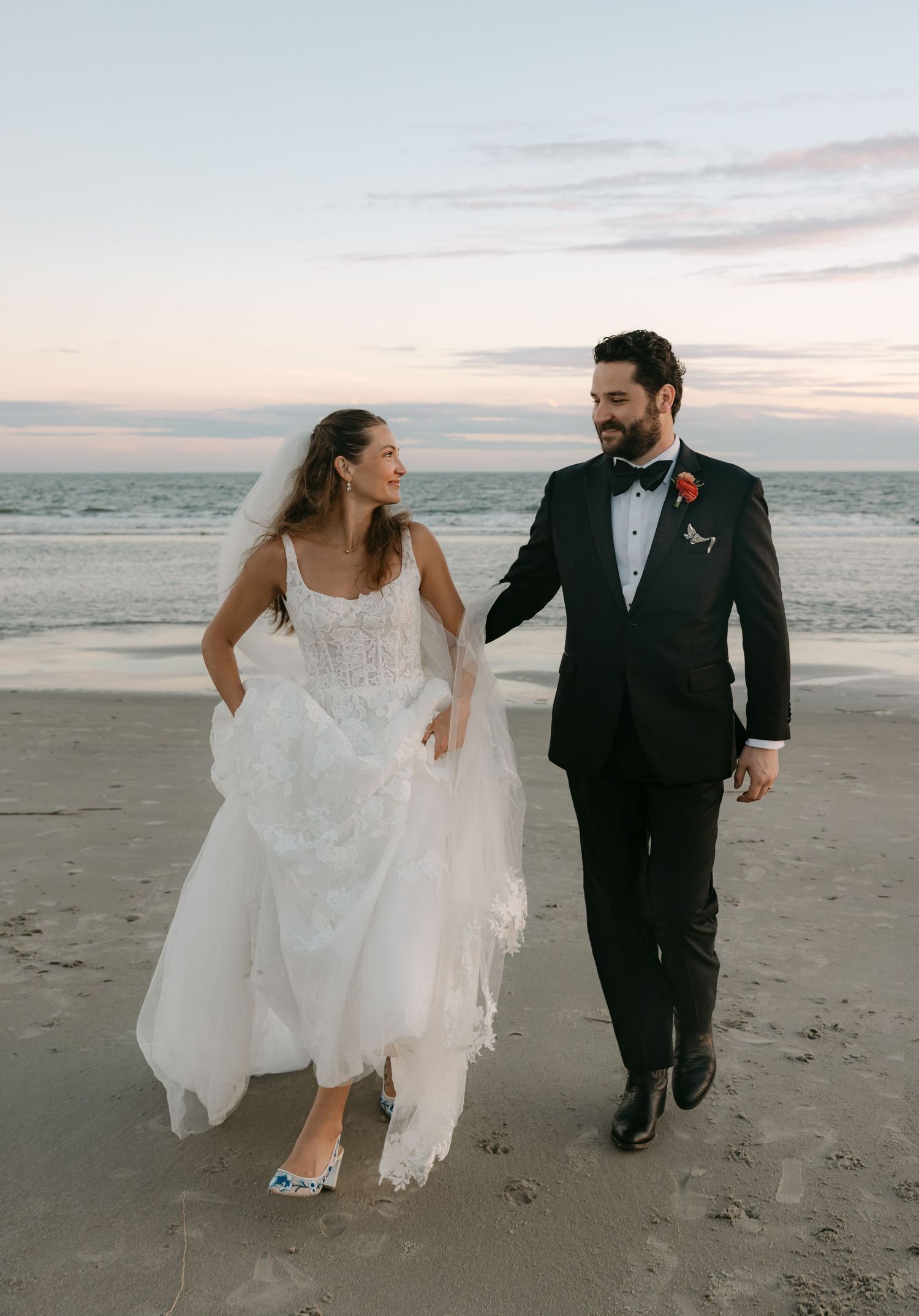 Bride and groom walk hand in hand on sandy beach at sunset in elegant wedding attire with ocean backdrop.